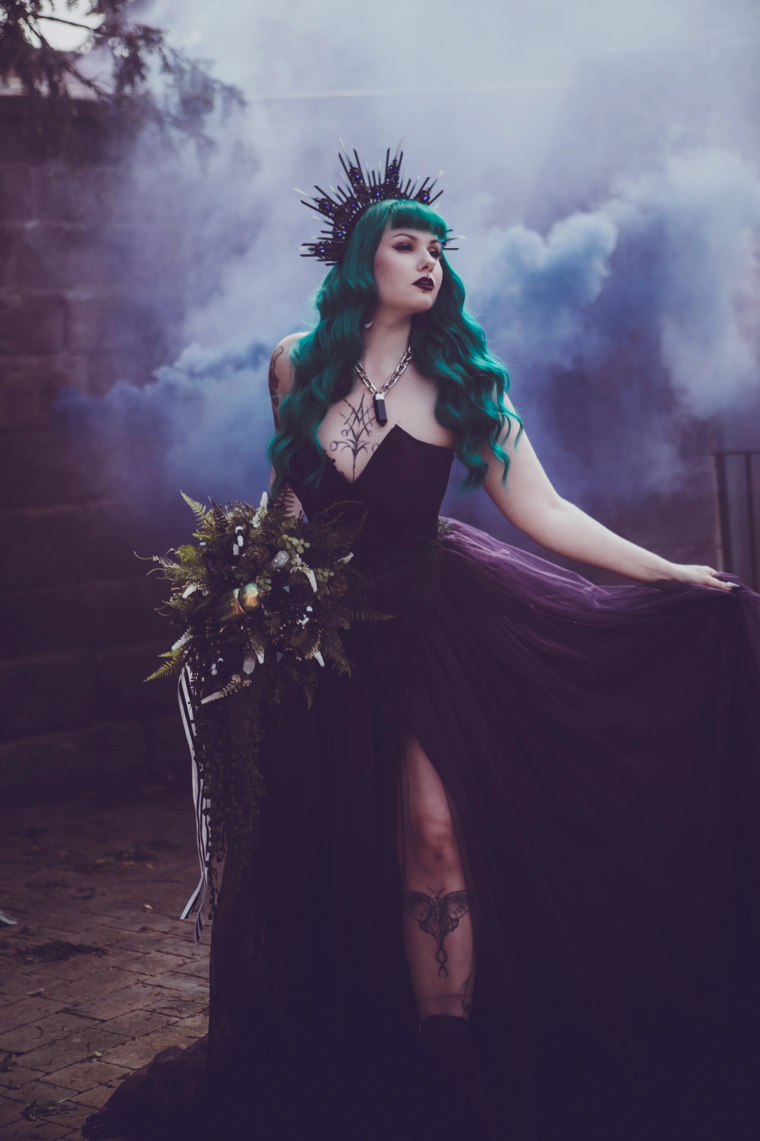 A woman with long teal hair and dark makeup, wearing a black strapless dress with a slit, stands outdoors in a foggy setting. She has a crown or headpiece with spikes, a large chain necklace, and tattoos on her chest and leg. She holds a dark bouquet of flowers and greenery.
