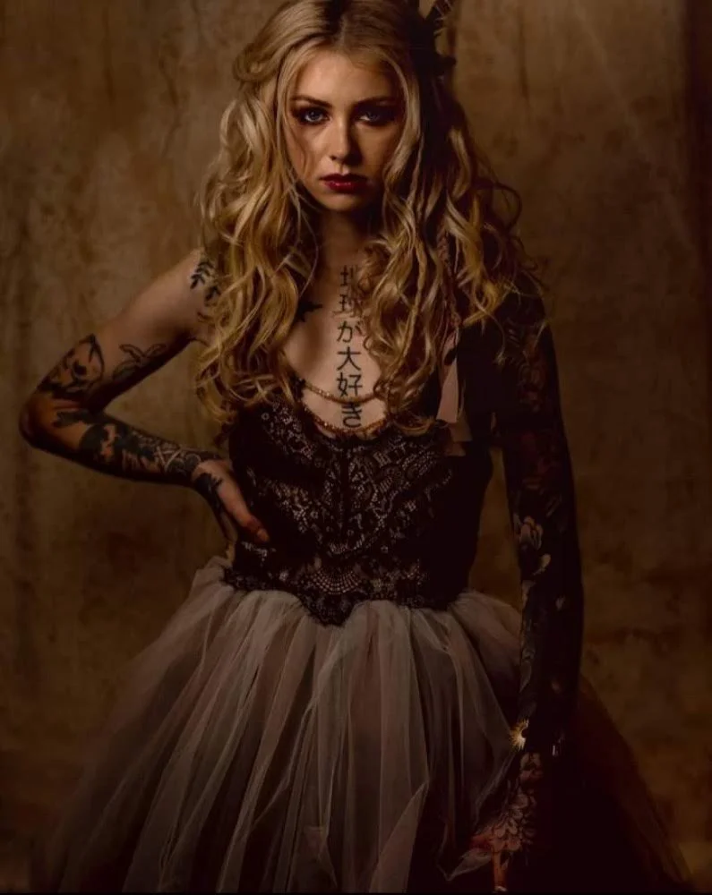 A woman with long, wavy blonde hair and tattoos on her arms and chest, wearing a black lace top and a tan tulle skirt, stands against a brown textured background with her hand on her hip and a serious expression.