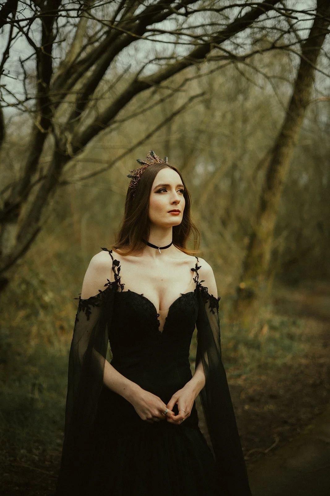 Forest Enchantress with pre-Raphaelite locks gazes softly into the depths of the forest. She wears a crown of green crystals and antique copper and a dress of darkest green, fully fitted bodice and sheer flowing sleeves. The dress is Hecate by Legend