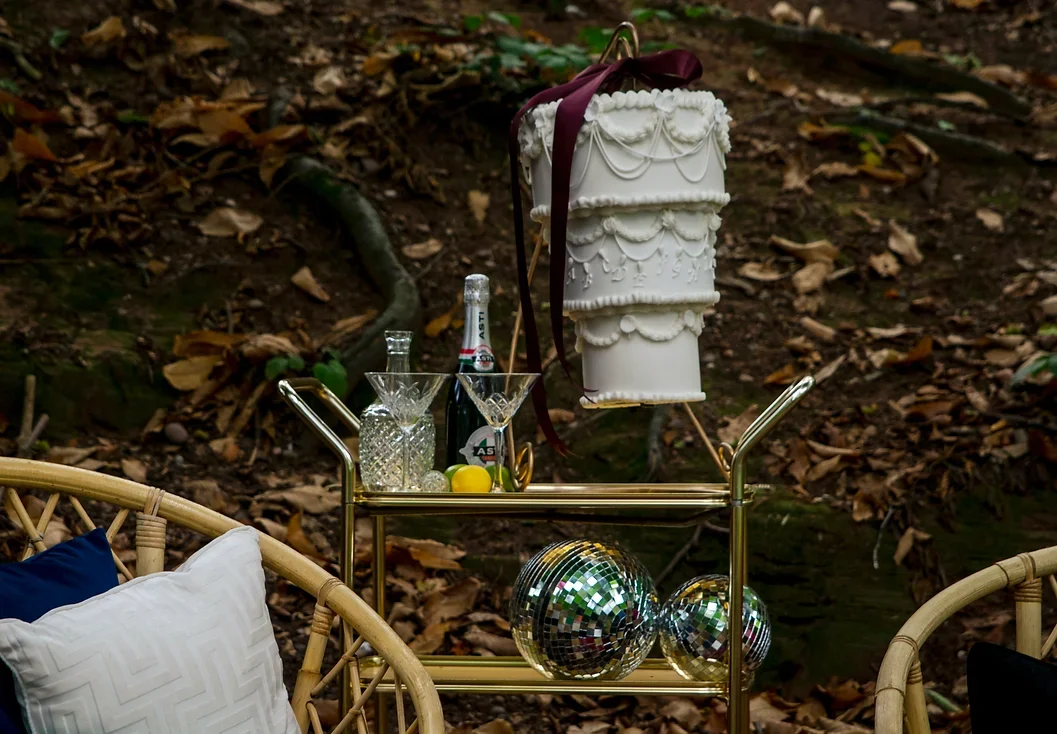 A decorative cake with white icing and ribbons placed on a bar cart in an outdoor setting with fall leaves, glassware, a bottle of champagne, lemons, and disco balls on the cart.