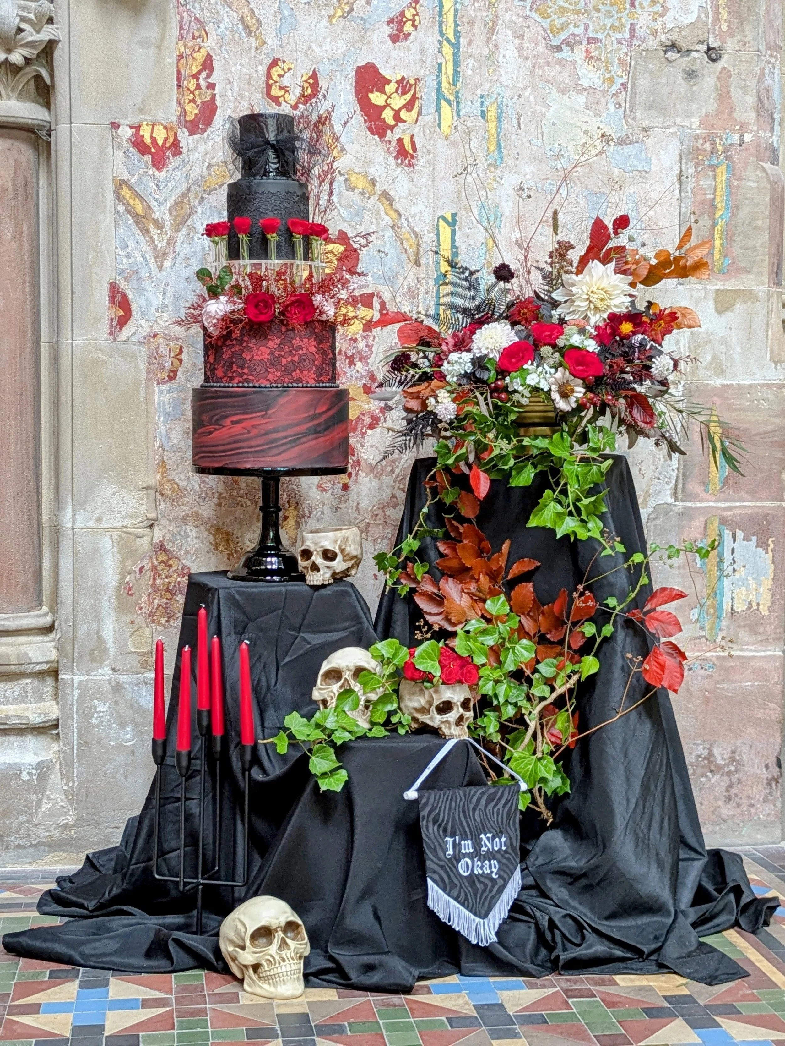 A Halloween-themed altar with skull decorations, red candles, and a large floral arrangement in a setting with an old, weathered wall.