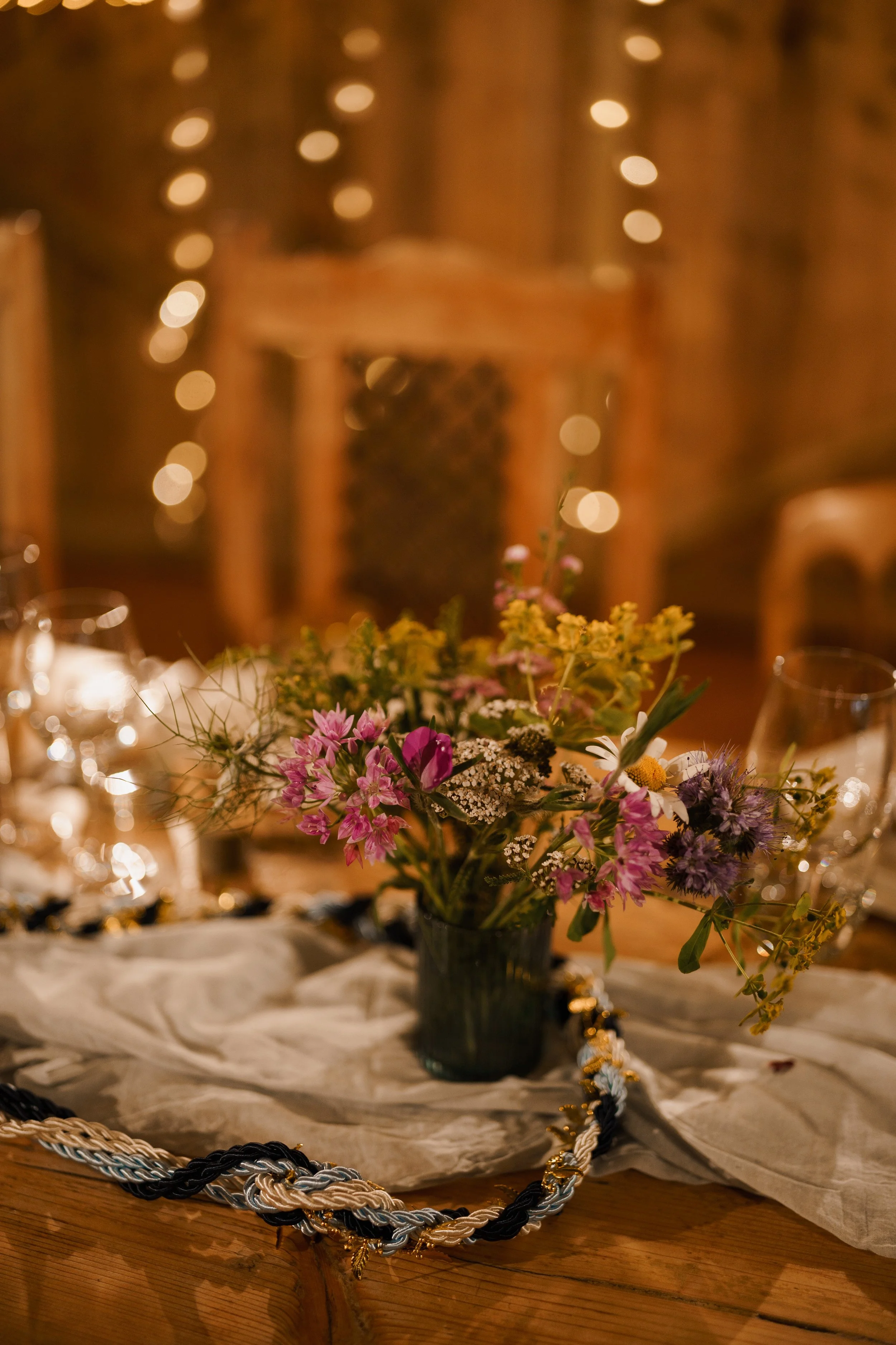A close-up of a table centerpiece with a small bouquet of flowers in a dark vase on a cream-colored fabric, with blurred background lights.