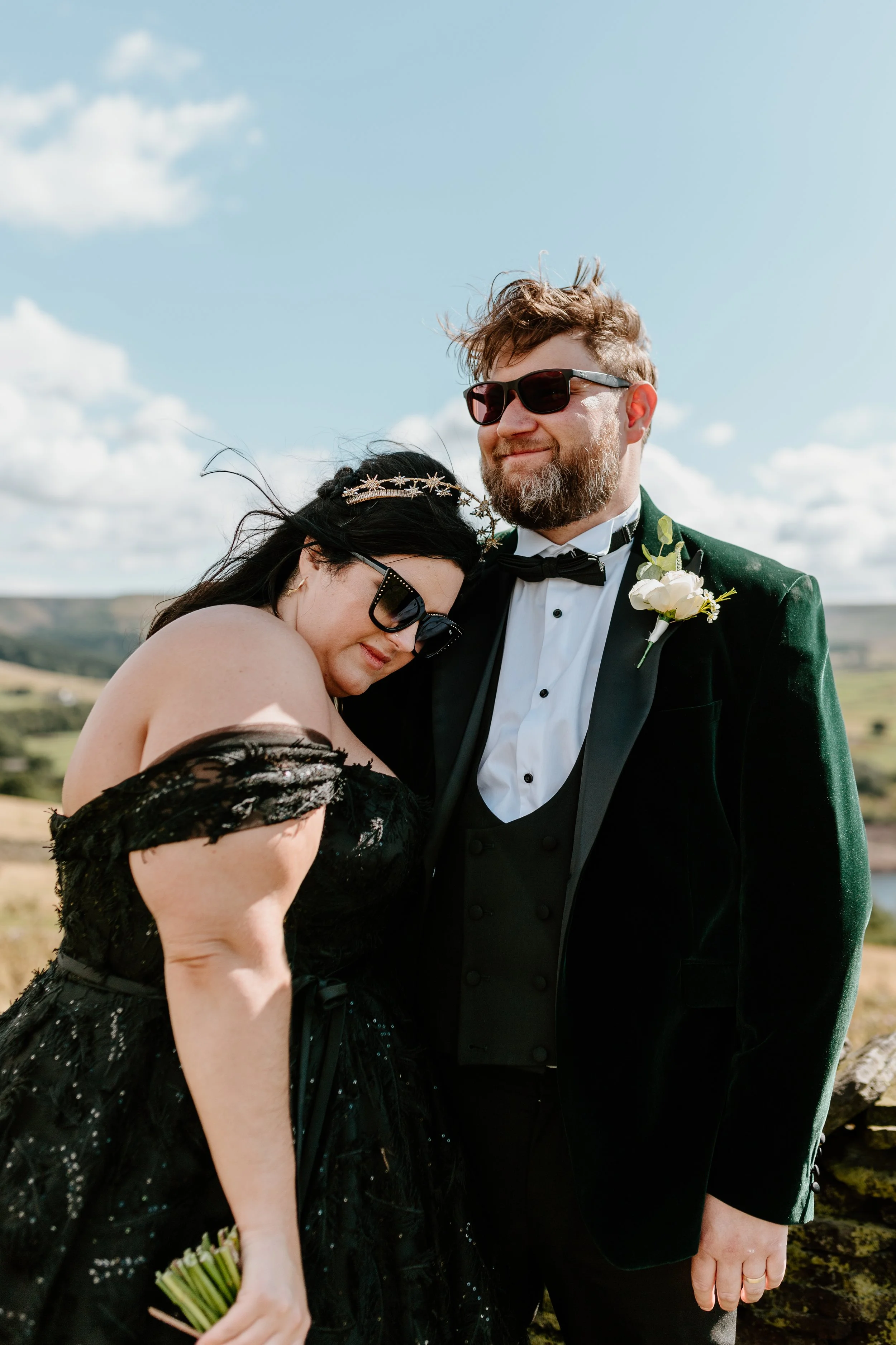 A bride and groom standing outdoors on their wedding day, with the bride resting her head on the groom's shoulder, both wearing sunglasses. The groom is dressed in a black tuxedo with a bow tie and boutonniere, while the bride wears an off-the-should