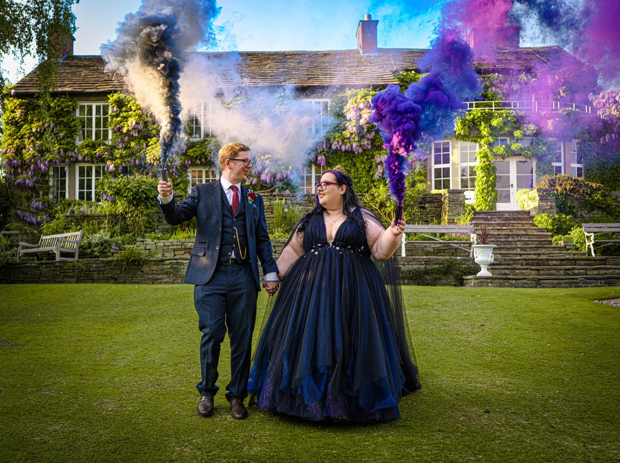 A couple dressed in wedding attire standing hand in hand on a lawn in front of a large house covered in climbing plants and flowers. They are holding smoke bombs emitting colorful smoke, one with white smoke and the other with purple smoke, and smili