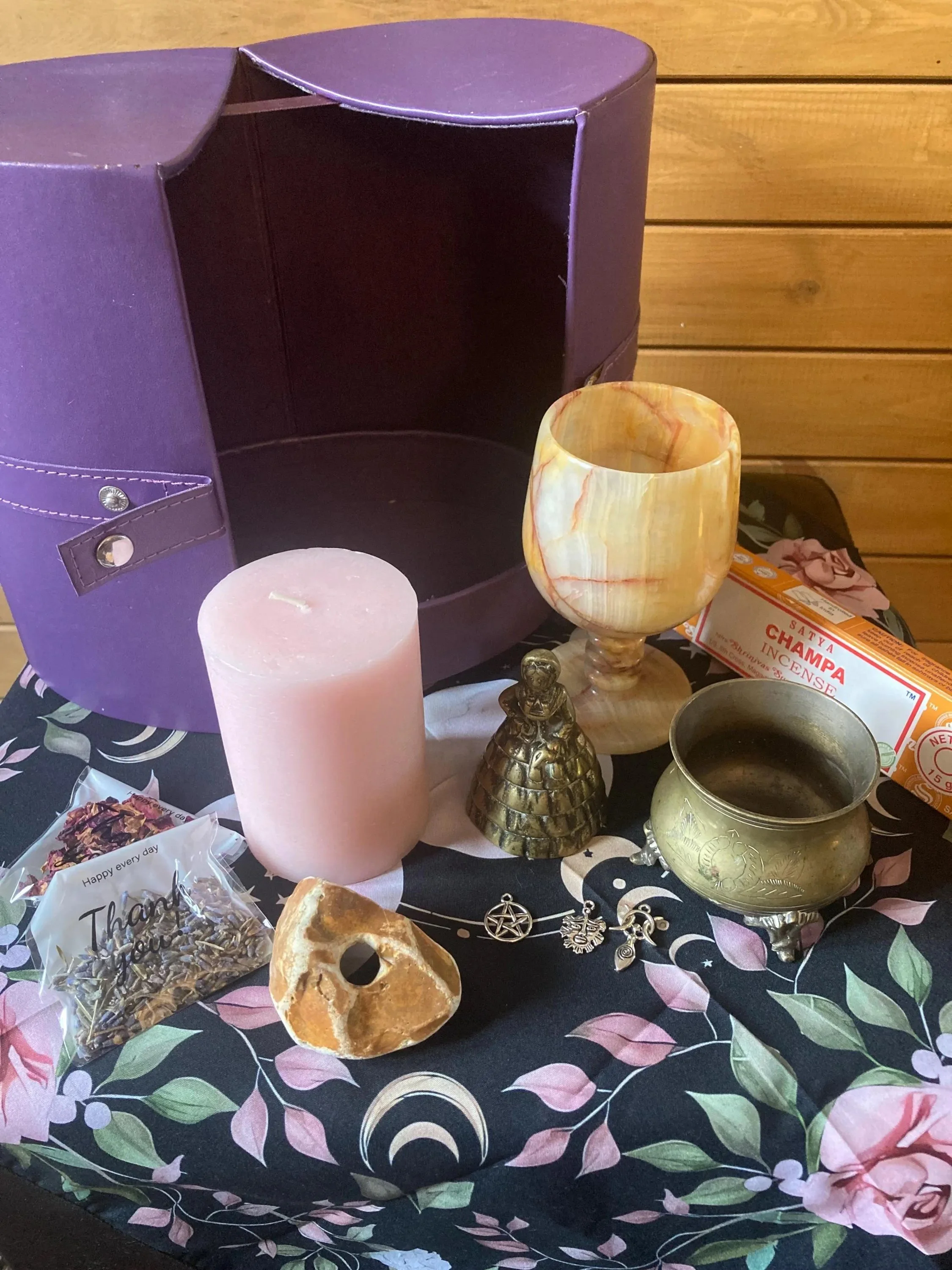 Collection of spiritual and decorative objects on a floral tablecloth, including a purple box, a pink candle, an orange and white marble goblet, a small golden figurine, a brass bowl, a pack of incense, a chunk of mineral, and a silver charm bracelet