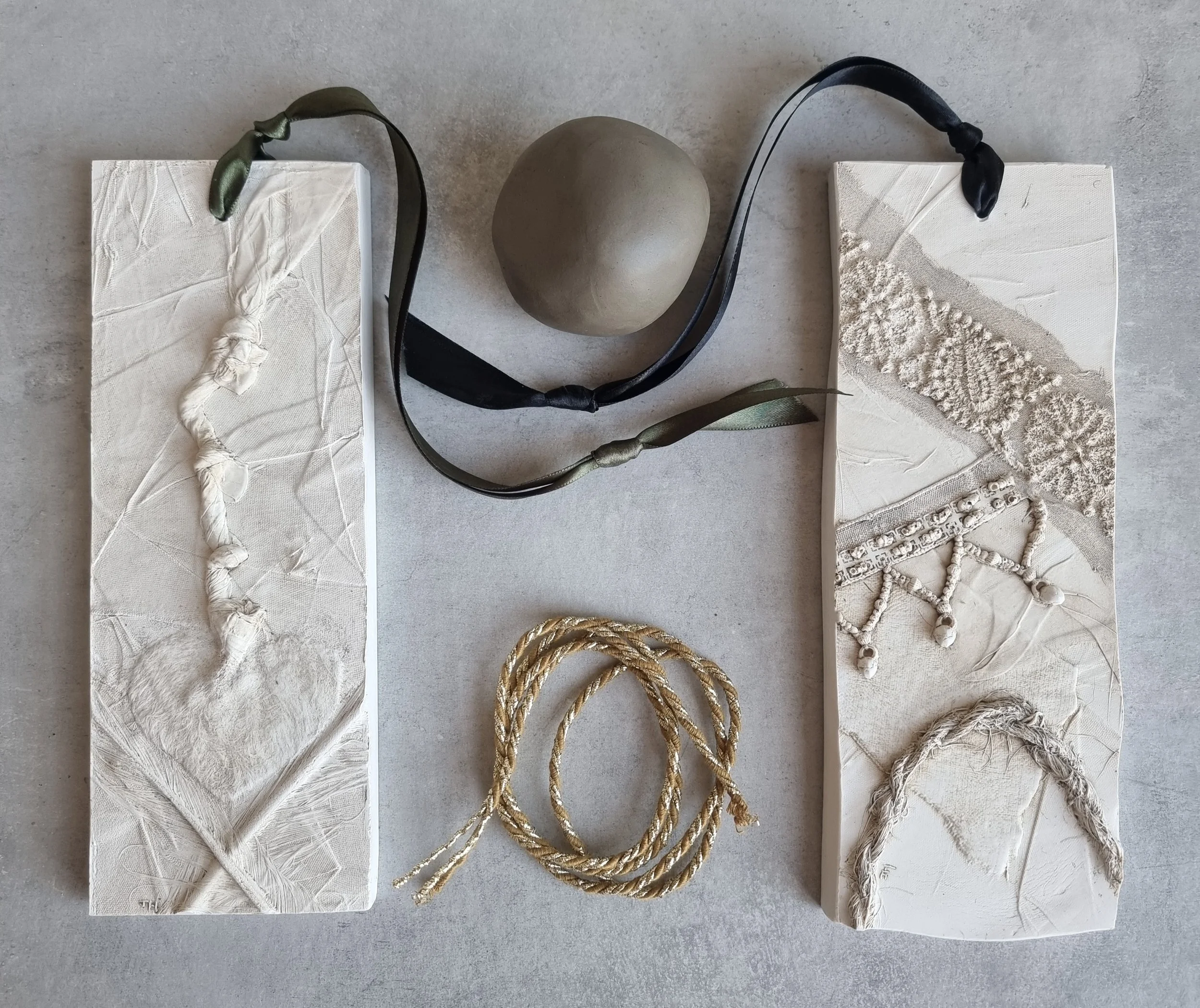 Two textured ceramic tiles with abstract designs, a small round gray stone, a black and olive green ribbon, and a gold braided cord arranged on a light-colored surface.