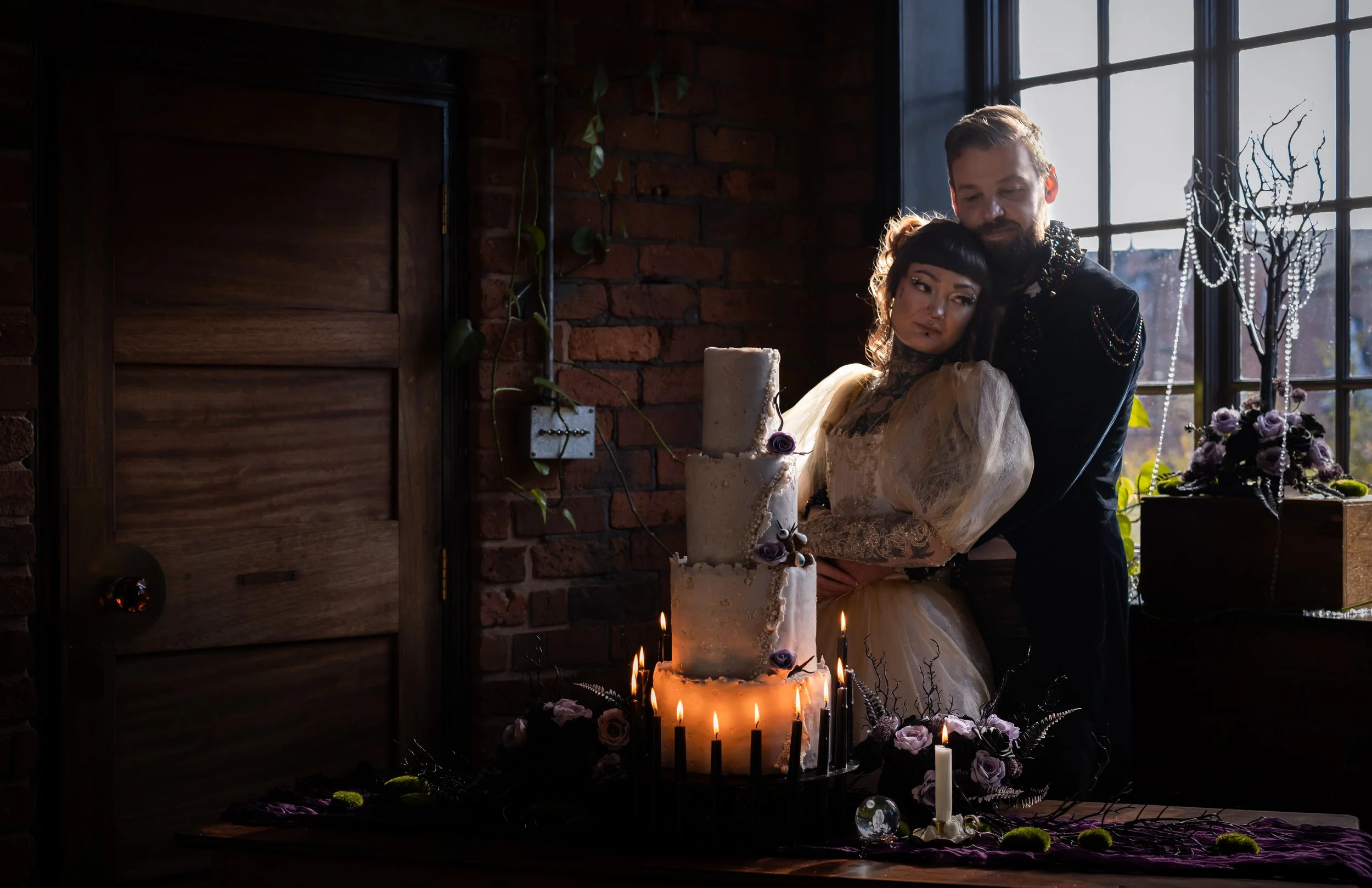 A couple stands close together behind a tall, lit cake with candles, decorated with purple flowers, in a dimly lit room with a brick wall, large window, and vintage decor.