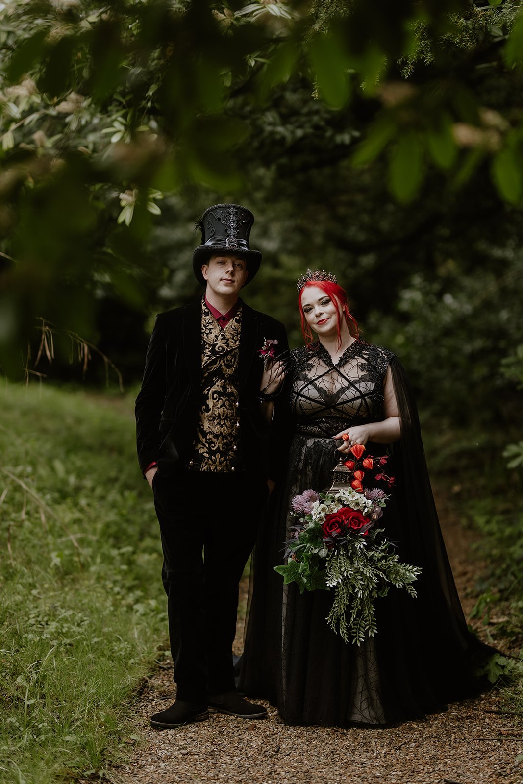 A man and woman dressed in gothic and Victorian style clothing standing outdoors on a dirt path surrounded by green foliage. The man wears a tall black top hat and a black velvet jacket with gold embroidery, and the woman wears a black lace gown, a t