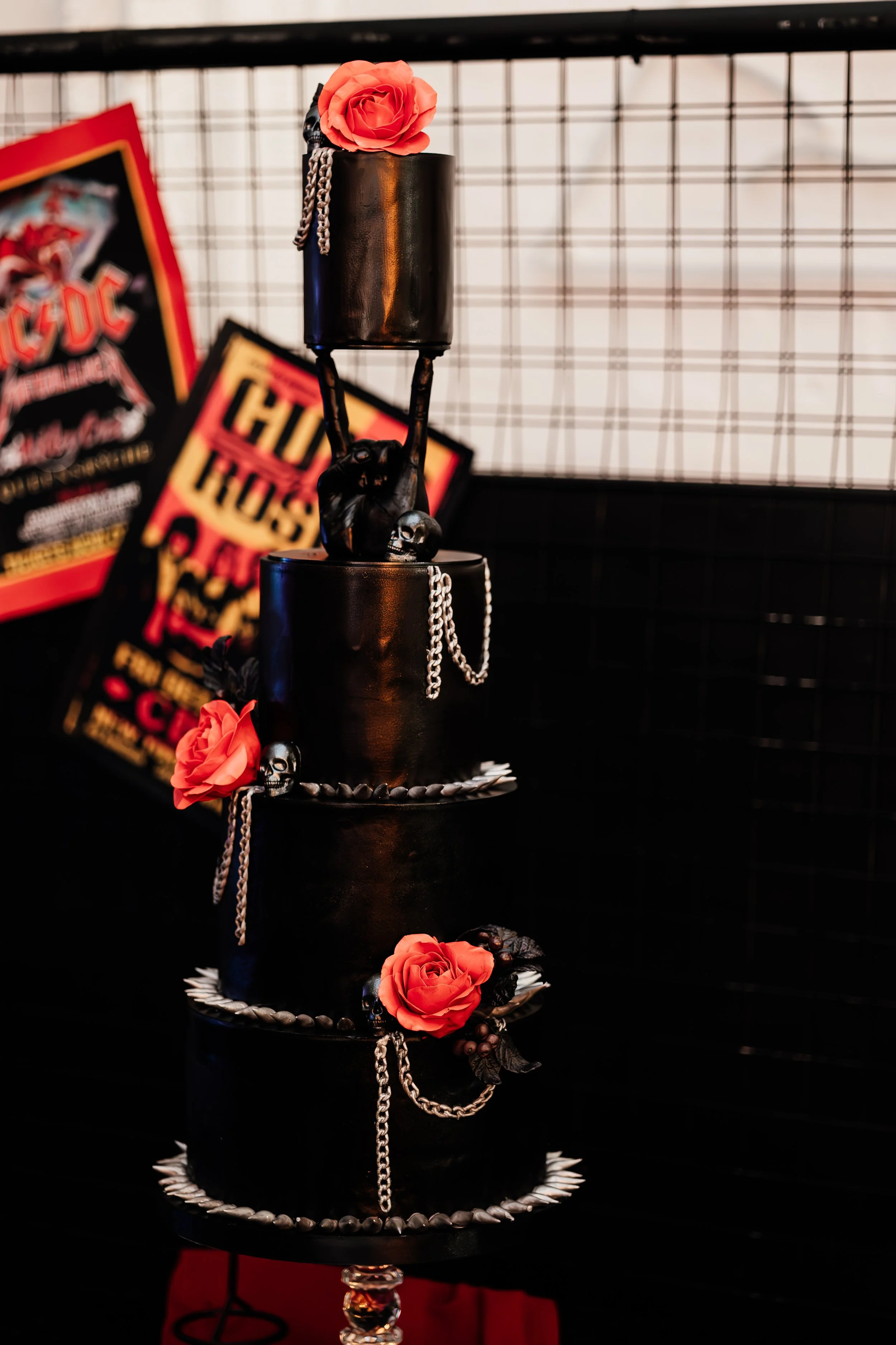 A black, four-tiered cake decorated with red roses, skulls, and silver chains. The cake is set against a black grid background with posters in the background.