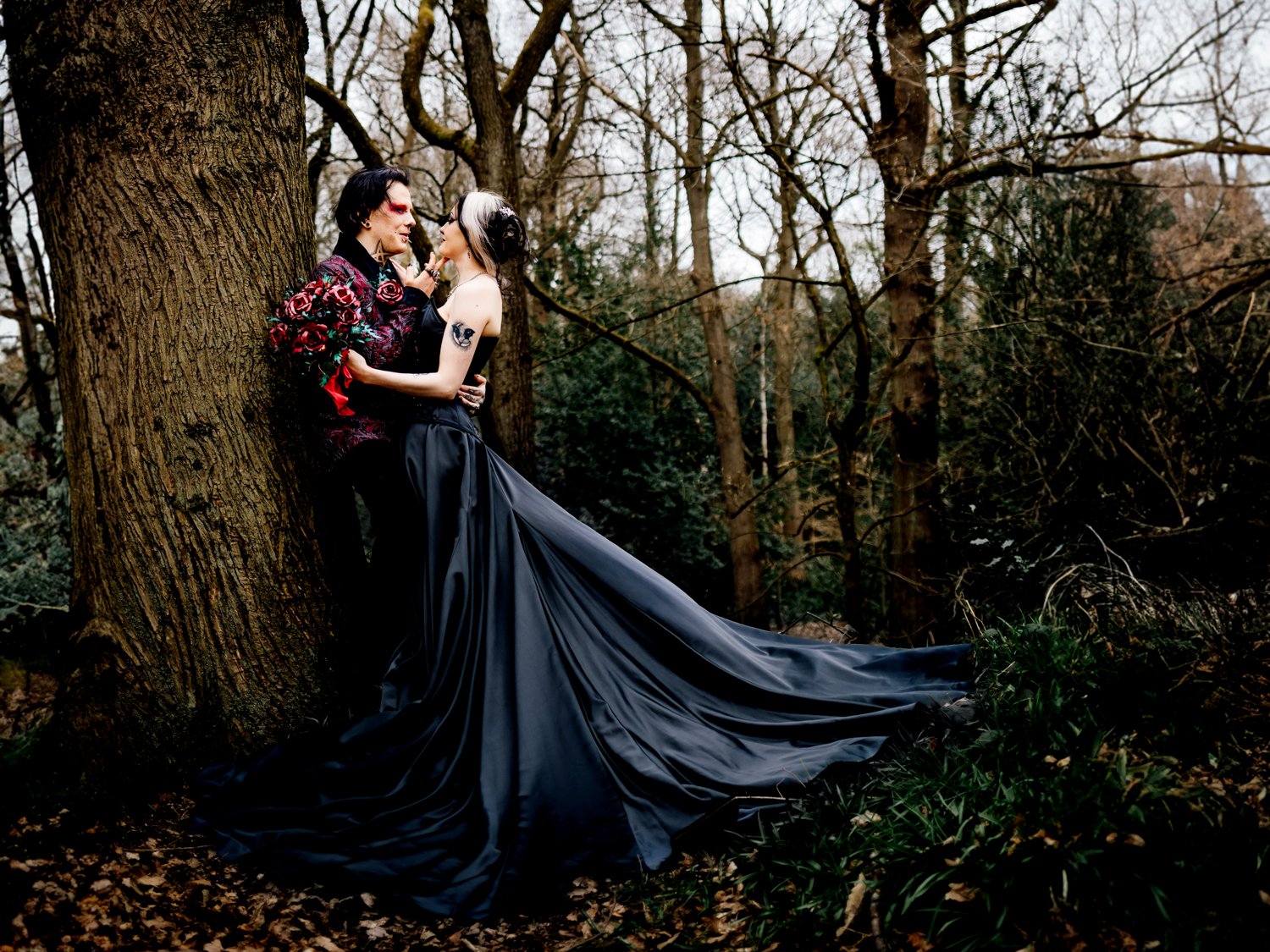 Two people dressed in gothic fashion standing close to each other against a tree in a forest with bare branches and dark foliage.