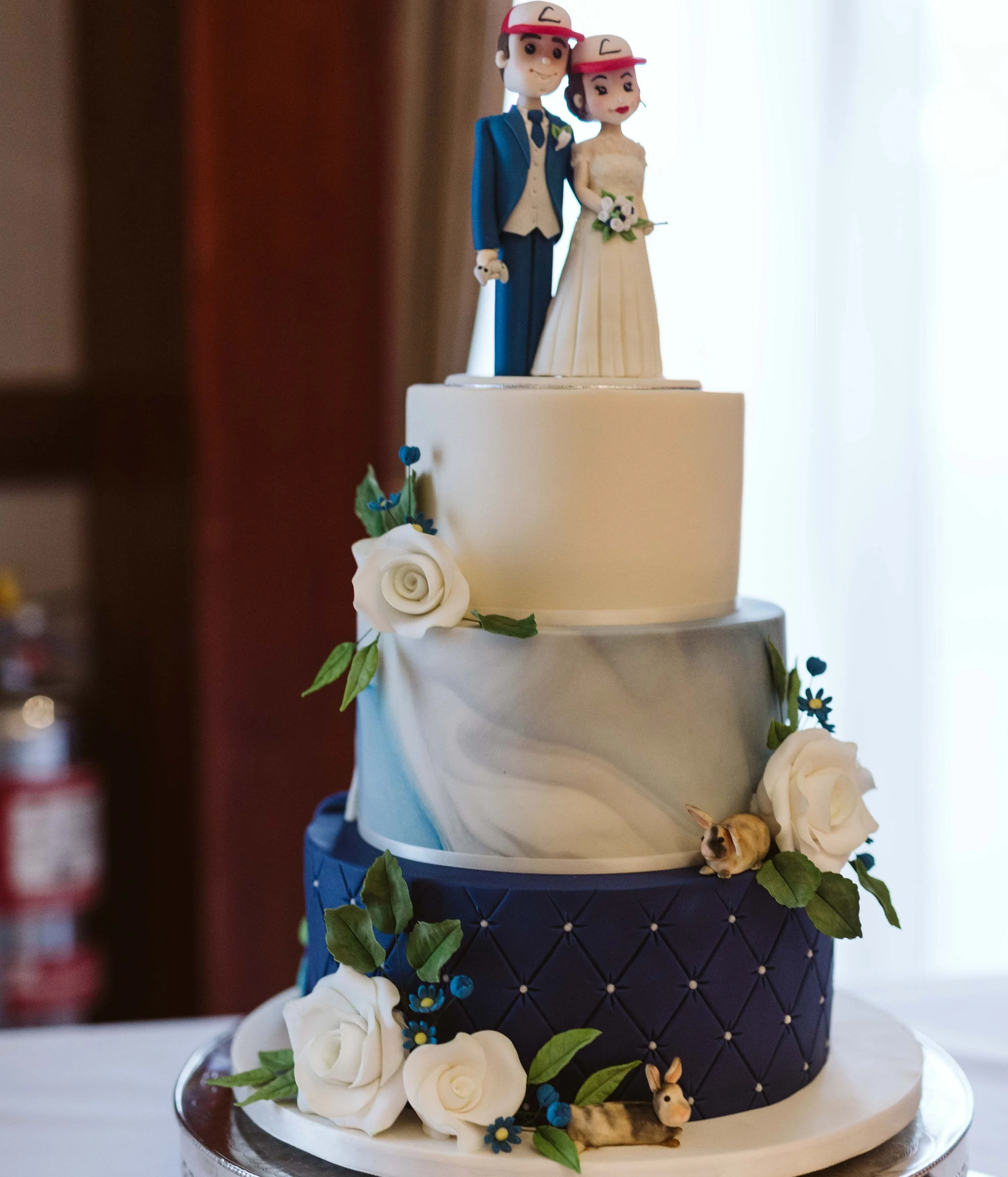 A multi-tier wedding cake decorated with white and blue fondant, white roses, greenery, and small animal figurines, topped with a bride and groom figurine wearing hats.