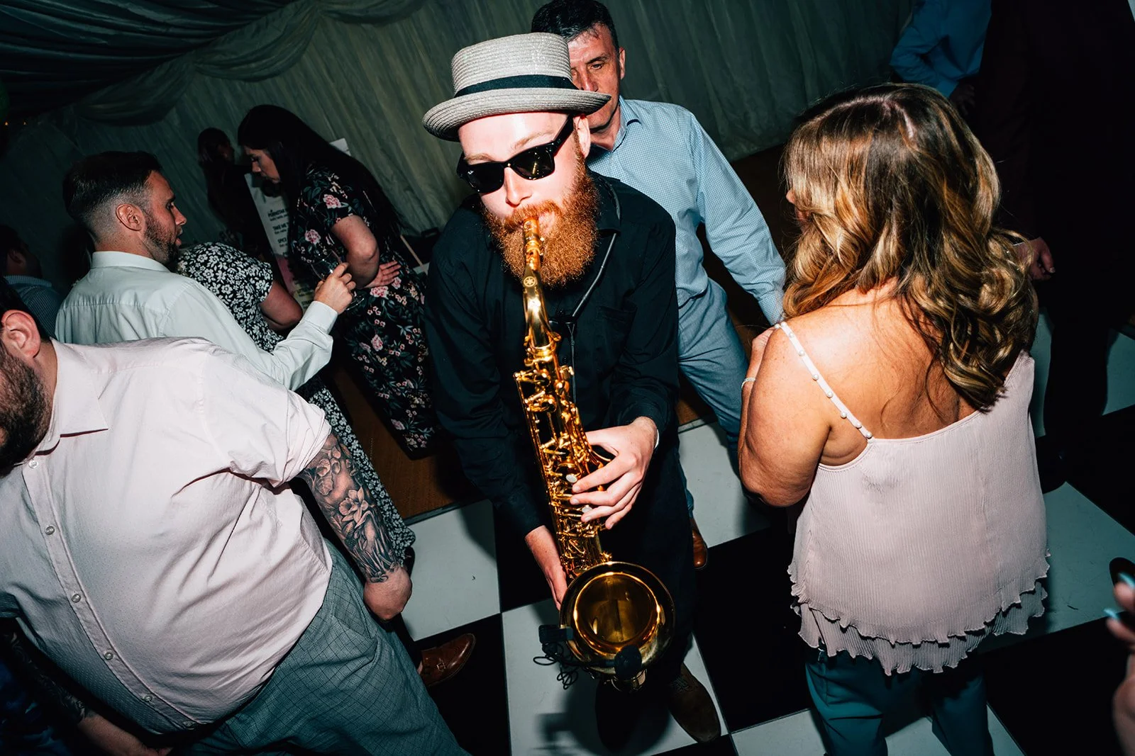 Man with a beard, sunglasses, hat, and black shirt playing a saxophone at a party surrounded by people dancing.