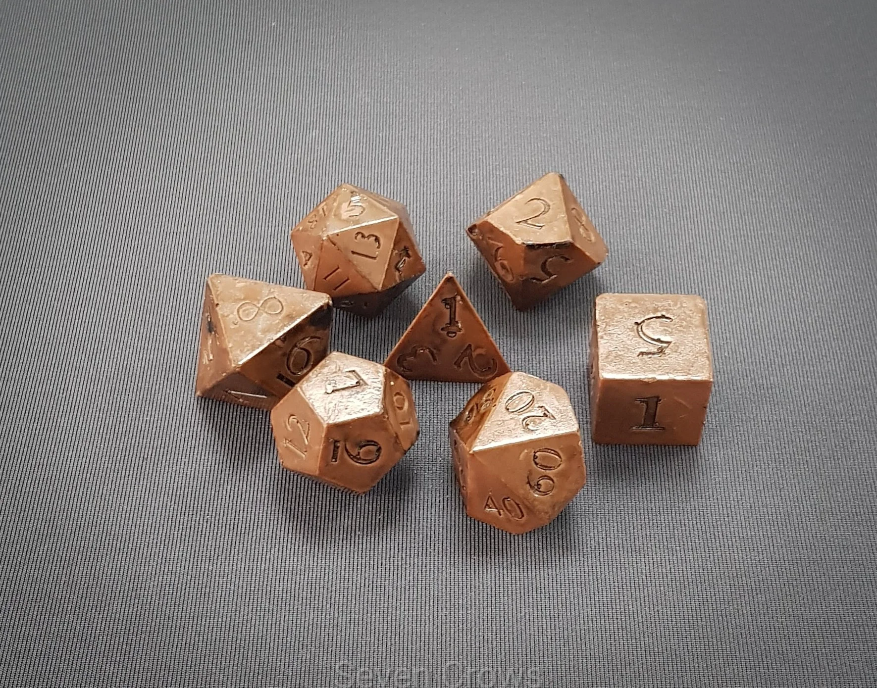 A collection of seven metallic polyhedral dice on a grey surface, including a four-sided, six-sided, eight-sided, ten-sided, and twenty-sided die, used for tabletop role-playing games.