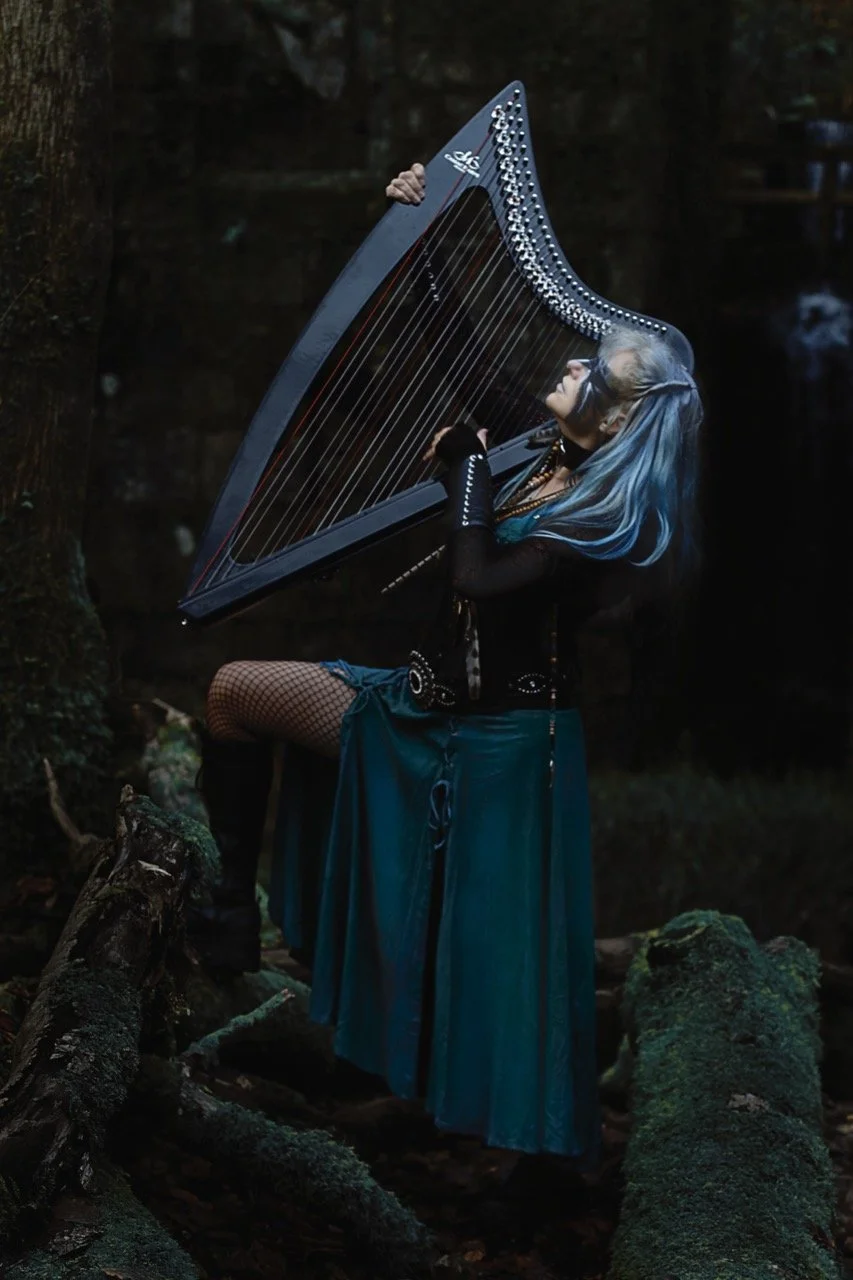 Person with blue hair playing a harp in a forest setting.