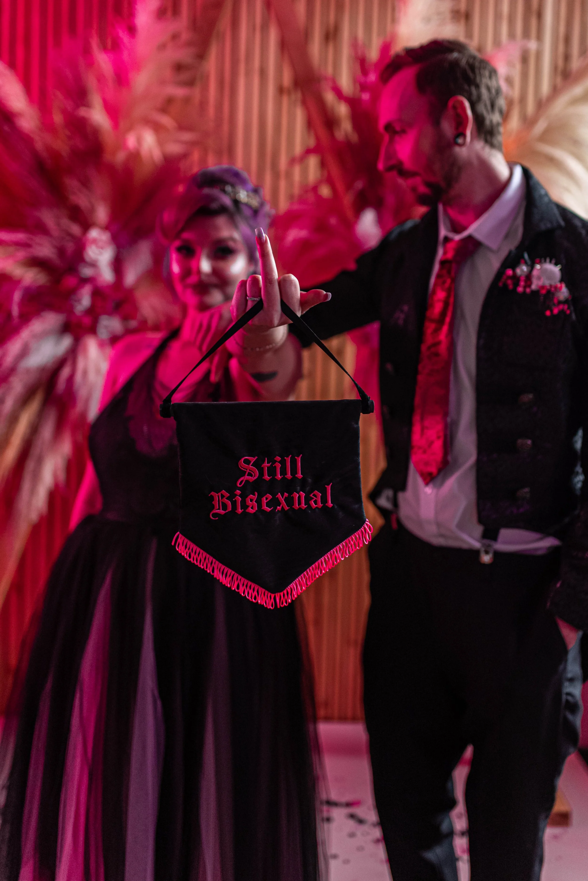 A couple holding a black flag with pink fringes that reads 'Still Bisexual' at a pink-lit event.
