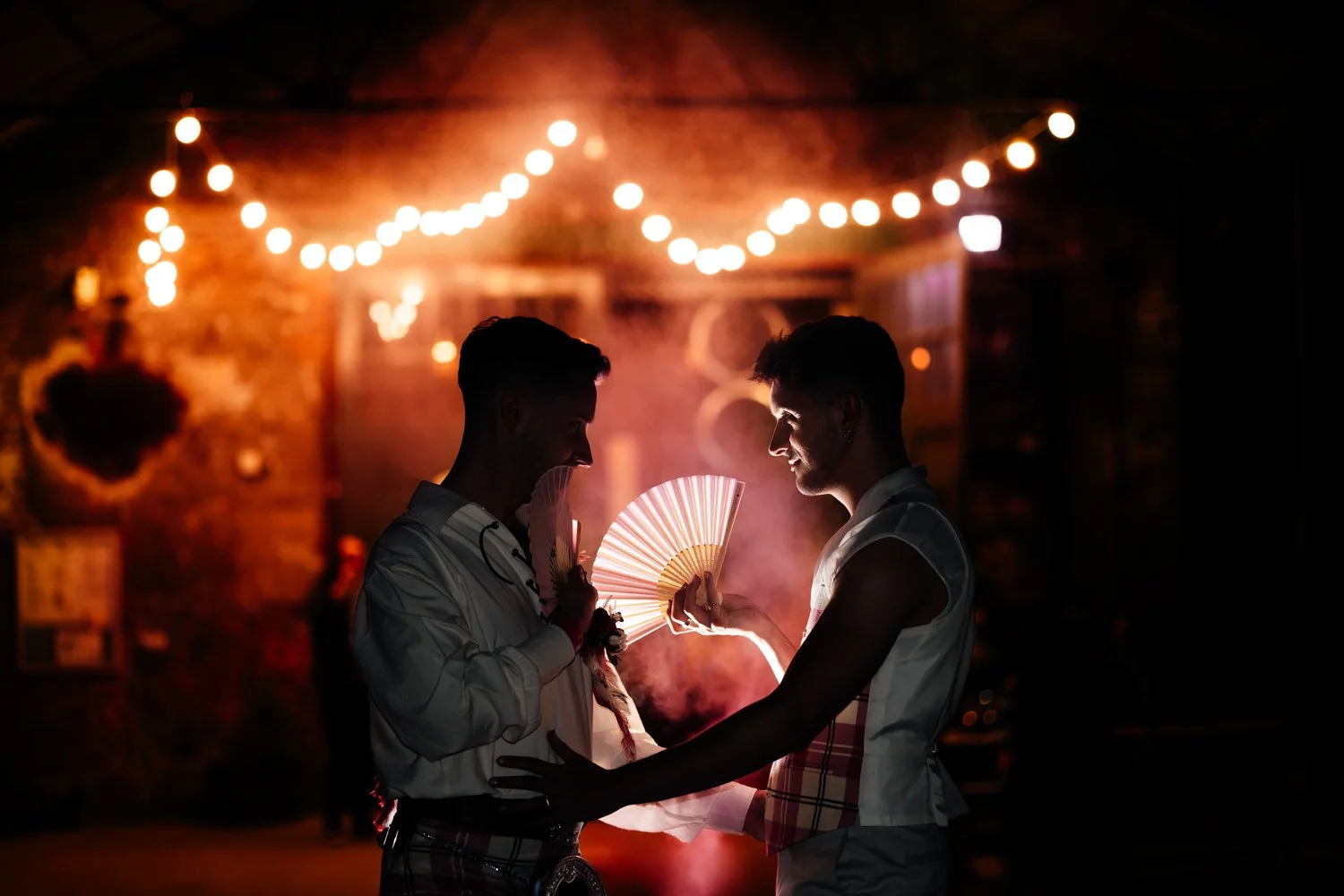 Two men smiling and holding fans face to face at a nighttime event with string lights overhead and a brick wall background.