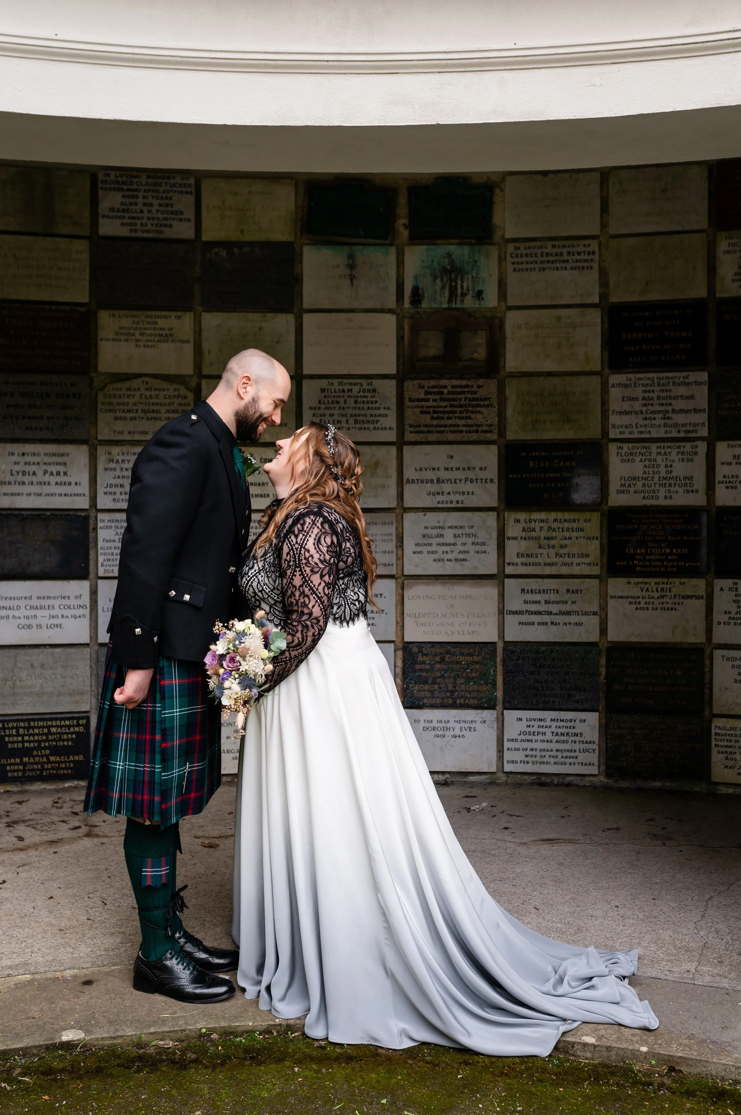 A newlywed couple is standing close together, gazing into each other's eyes, outside at a memorial wall. The groom is wearing a traditional Scottish kilt and a black jacket. The bride is wearing a white gown with a lace top and a black lace shawl, ho