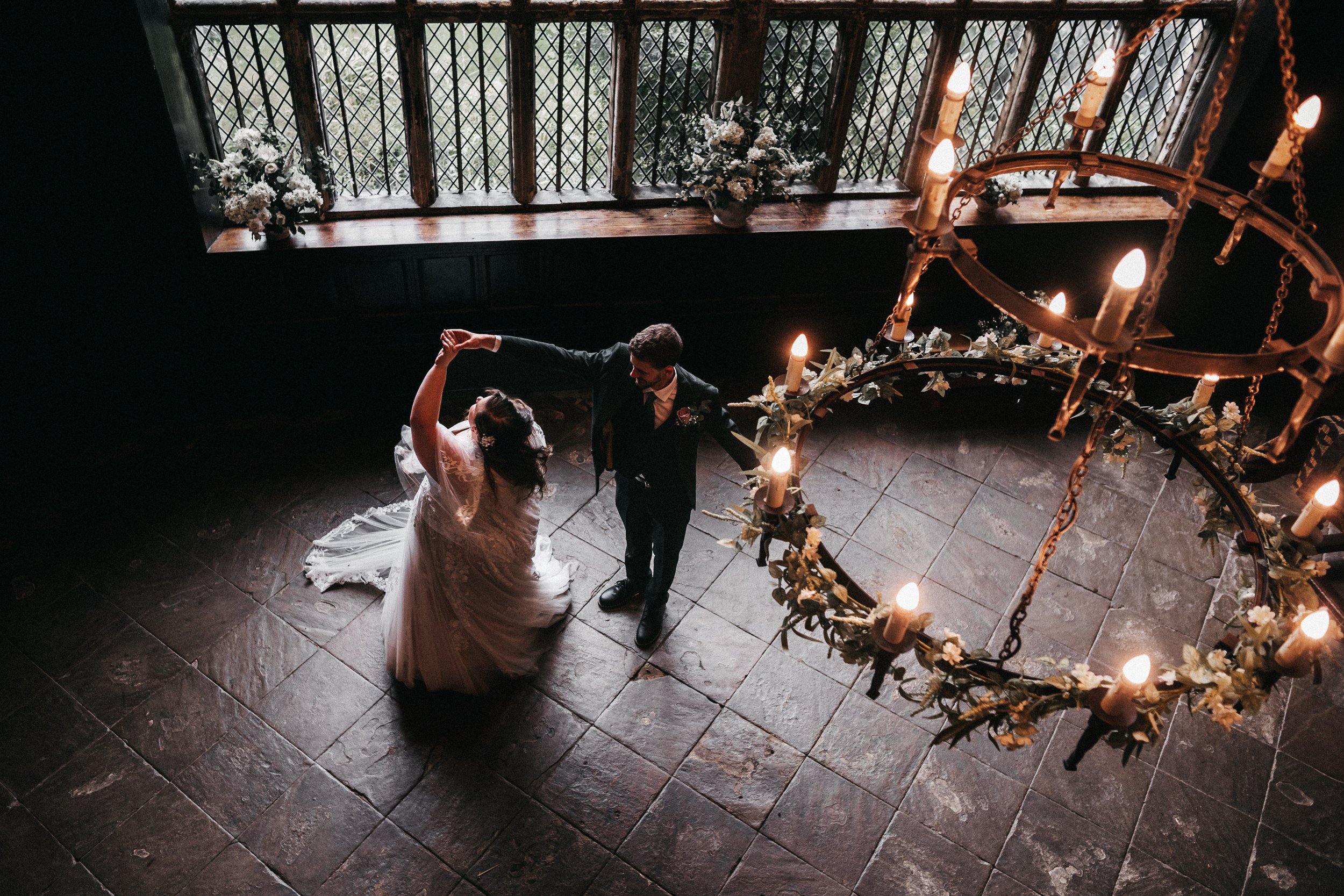 A bride and groom dancing in a dimly lit room with large windows, a chandelier hanging overhead, and floral arrangements on the window sill.