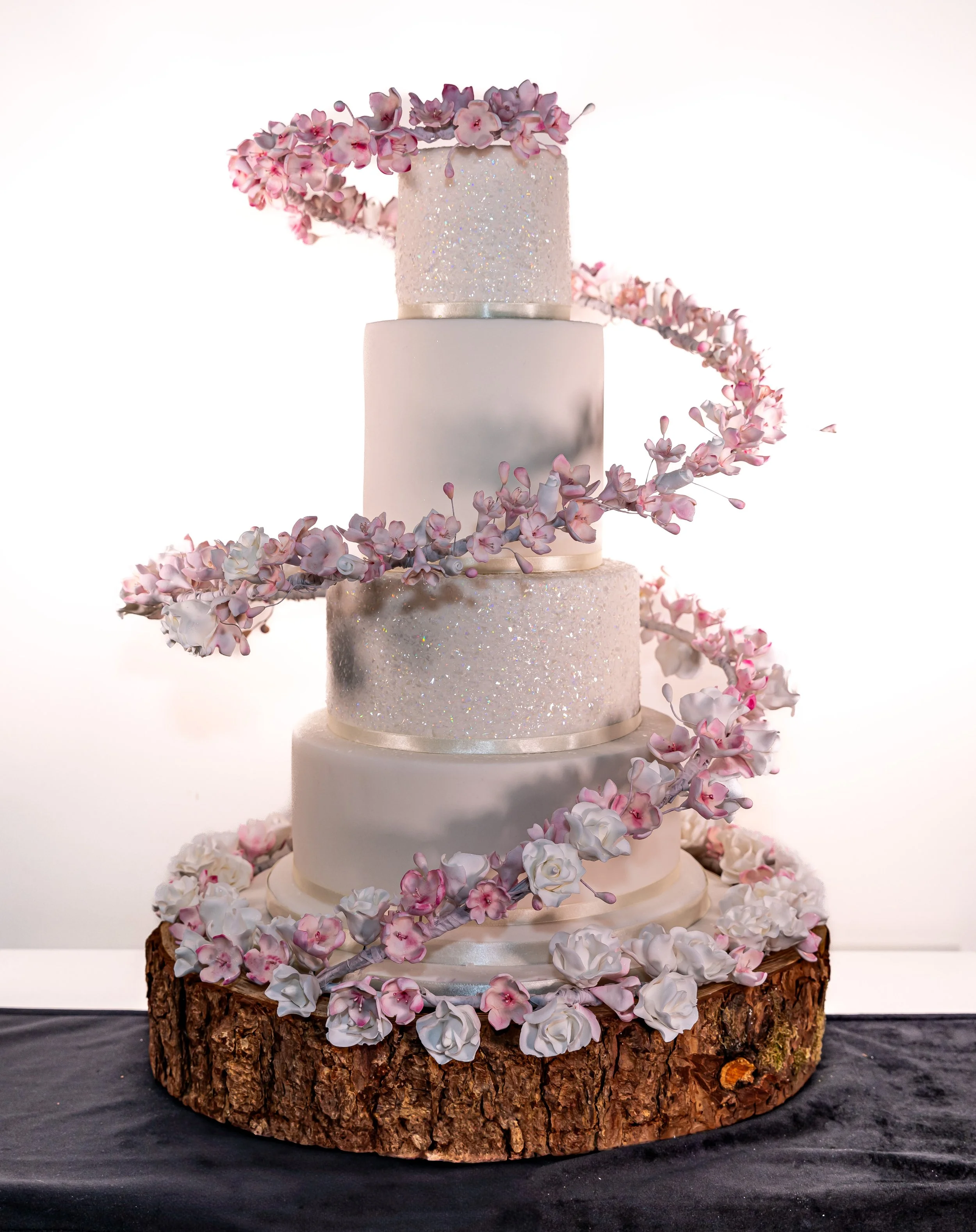 Four-tiered wedding cake on a wooden base decorated with pink and white flowers, with a spiraling floral decoration.