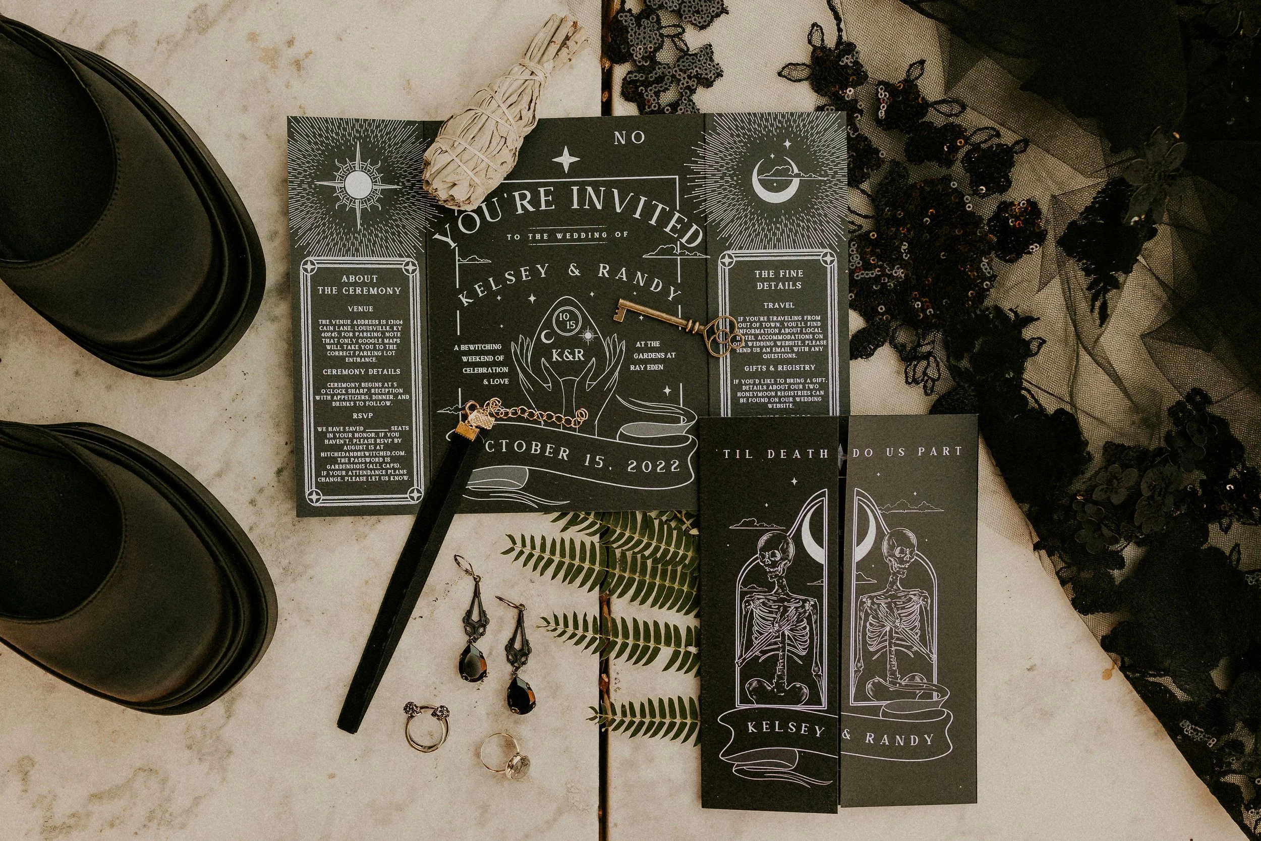 Black wedding invitation with white text and celestial illustrations, featuring a key, ferns, jewelry, black shoes, a rolled bundle of sage, black lace, and sequin decorations.
