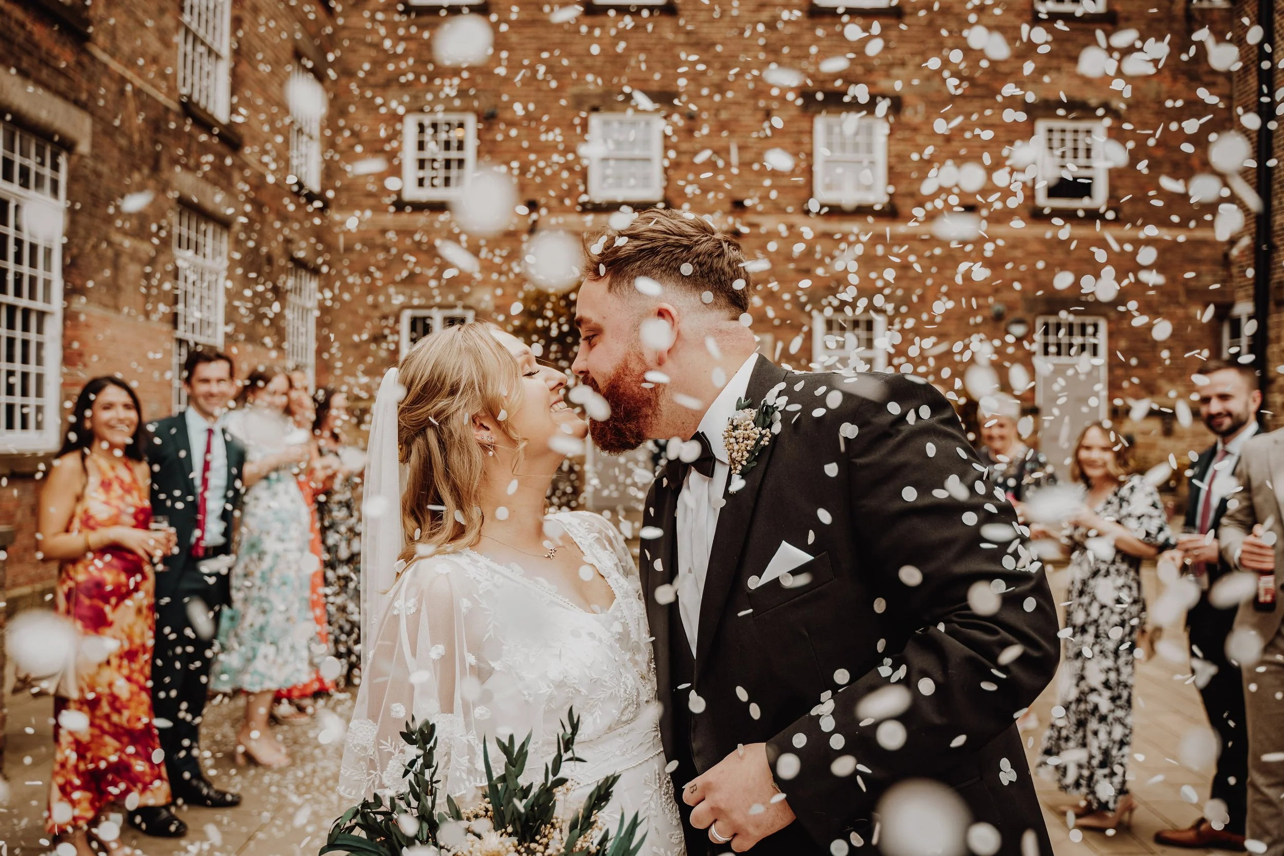 A bride and groom share a kiss during their wedding celebration as confetti falls around them, with guests cheering in the background at an outdoor brick courtyard.
