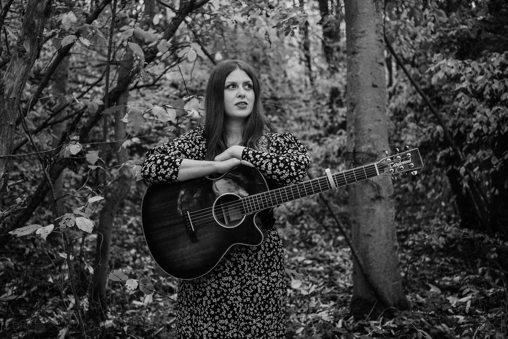 A woman with long hair holding a guitar, standing in a wooded area surrounded by trees and leaves.