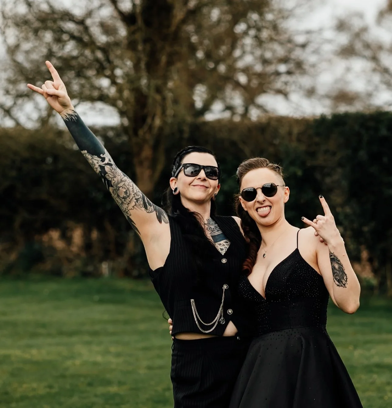 Two women posing outdoors with trees in the background, making rock and roll hand signs and smiling.