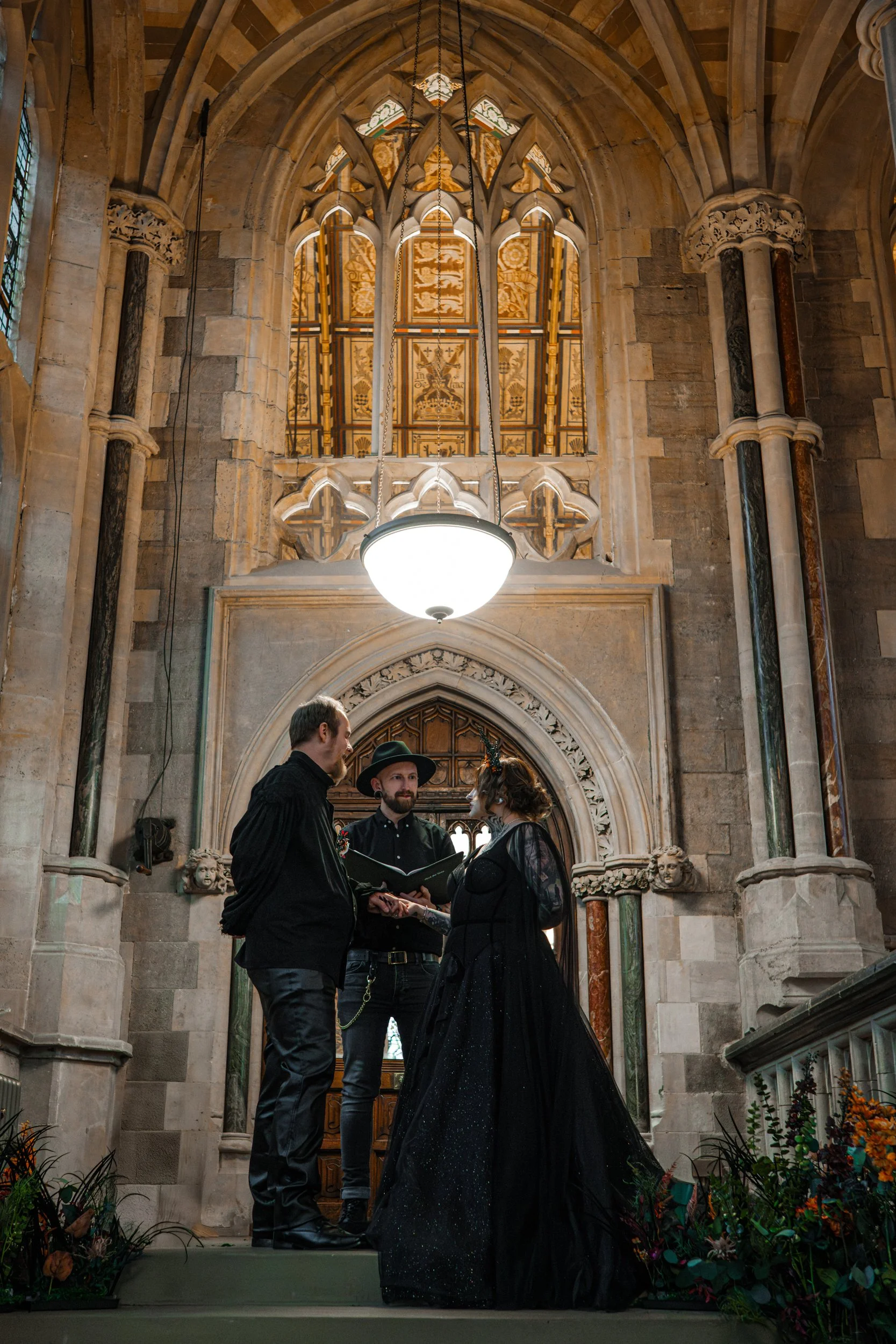 A wedding ceremony inside a historic church, with a couple in black wedding attire standing before an officiant, who reads from a book, and behind a large stained-glass window.