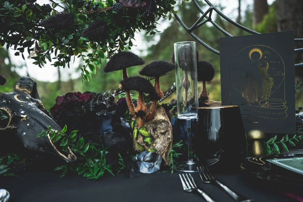 An outdoor table decorated with gothic-themed items, including a skull and mushrooms, surrounded by greenery, with a black tablecloth, glassware, and a card featuring celestial imagery.