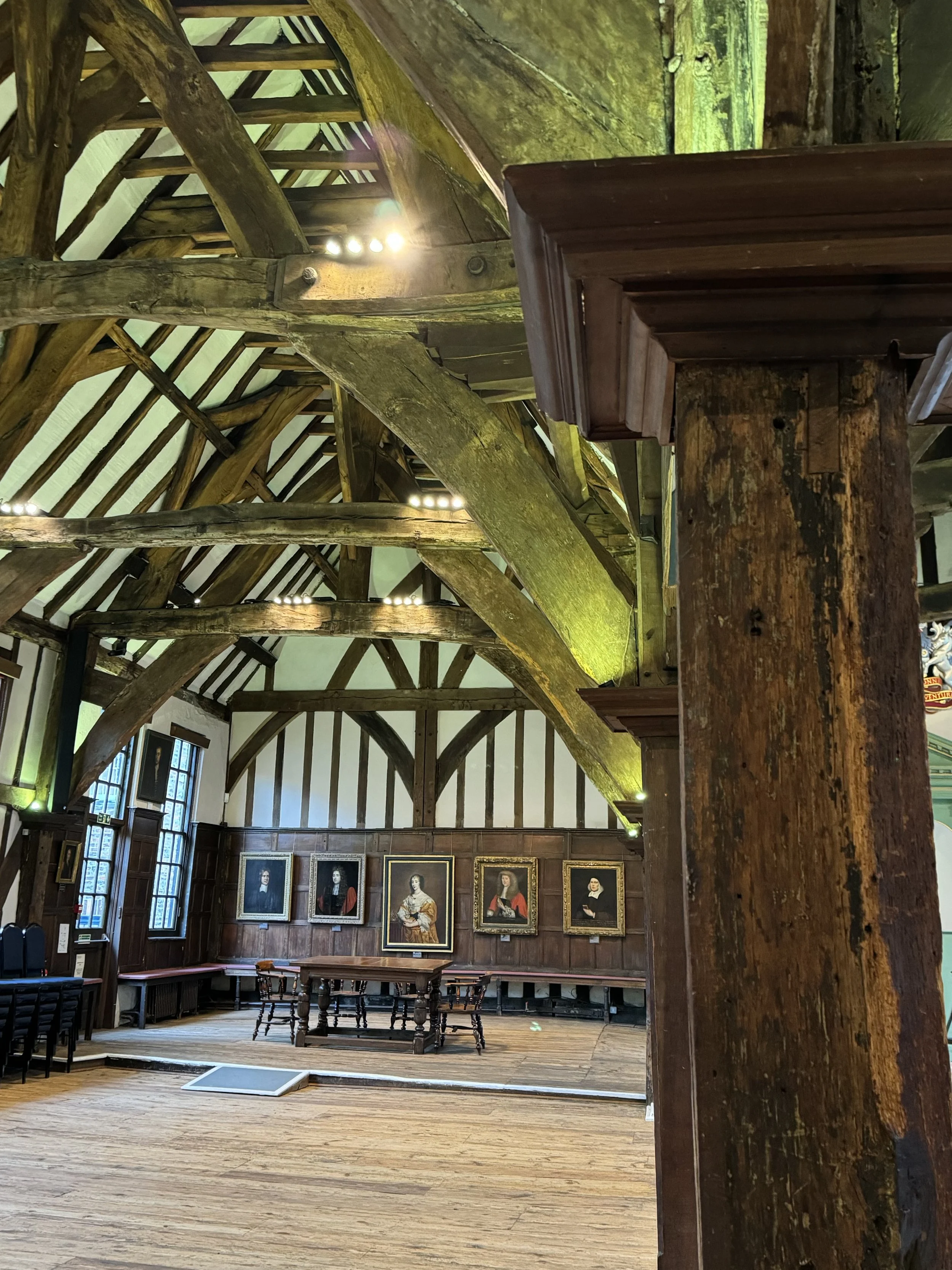 Interior of a historic building with wooden beams, portraits on the wall, and a wooden table with chairs.