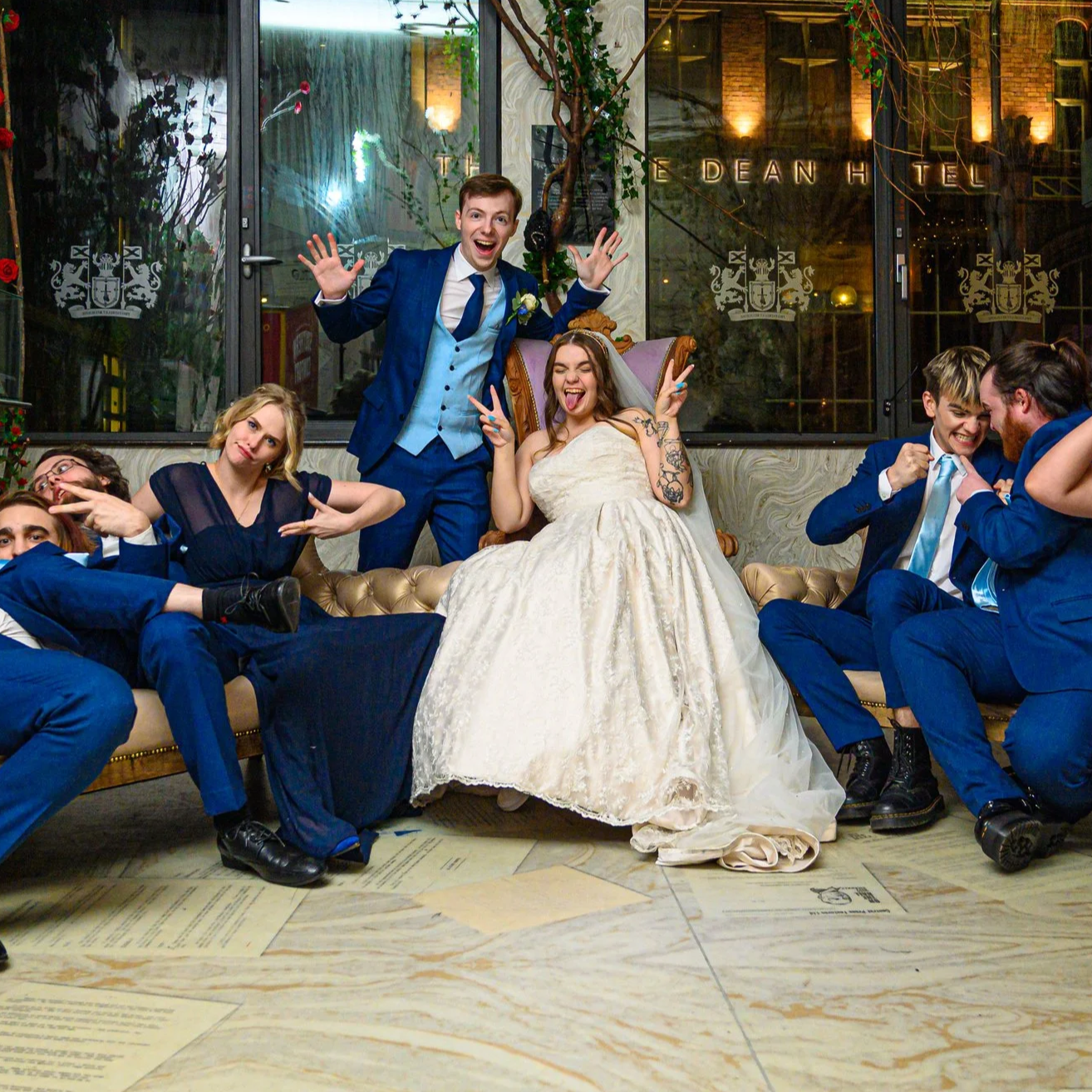 Group of people at a wedding celebration, with the bride and groom sitting on a couch, all making playful gestures and smiling in a lively indoor setting.