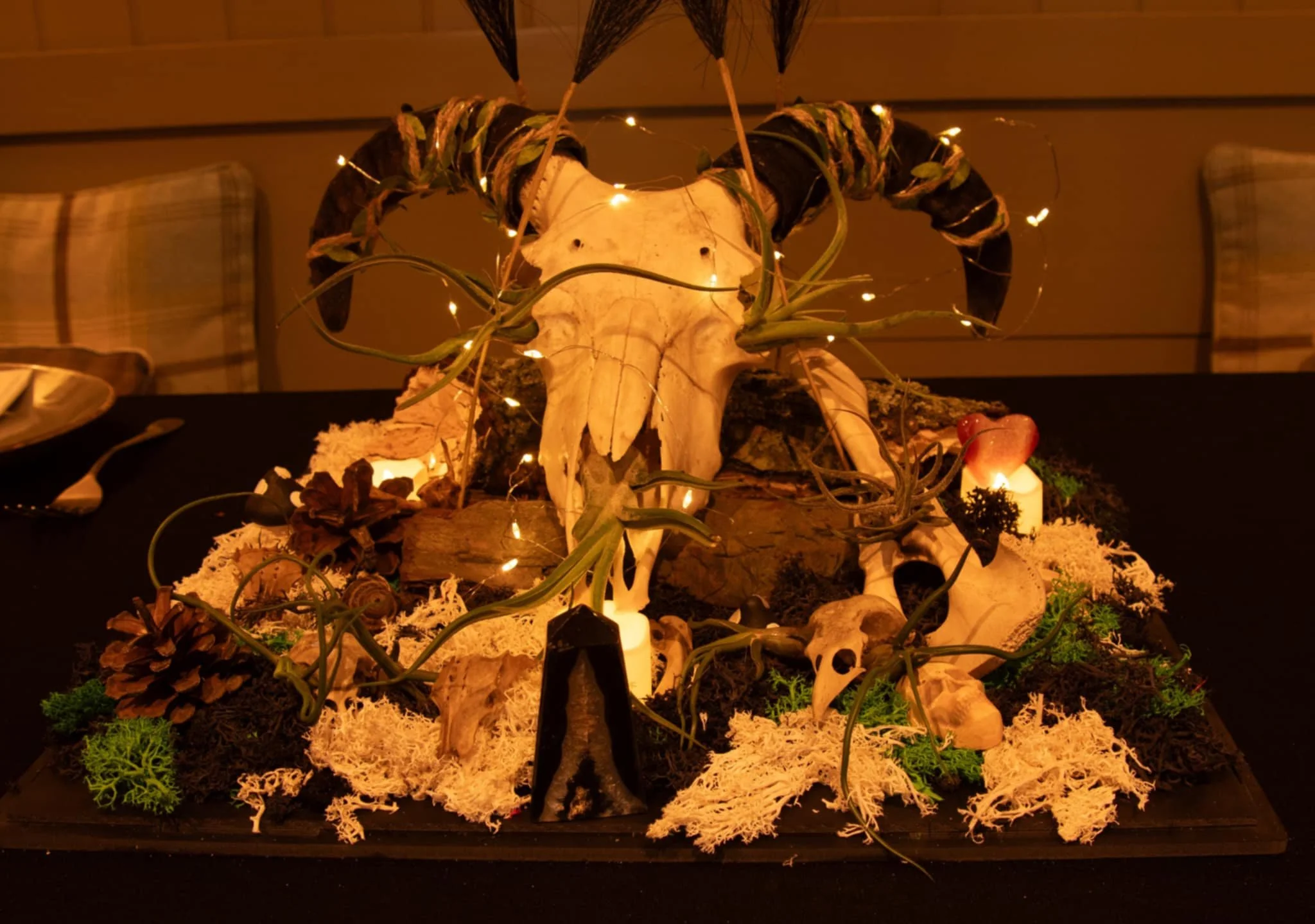A decorative centerpiece featuring a large animal skull with horns, surrounded by greenery, moss, pine cones, and small lights on a table at a dimly lit setting.