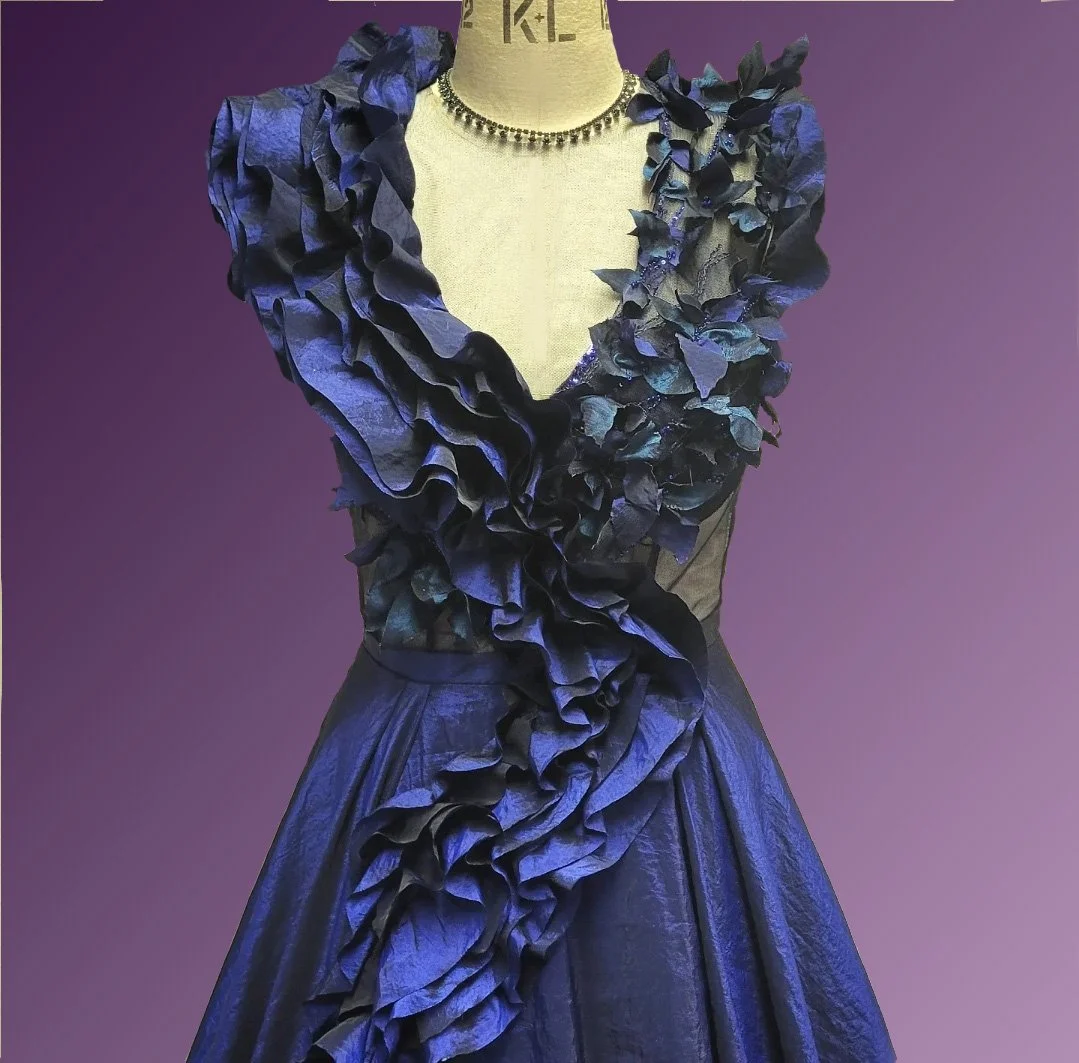 A formal evening gown in navy blue with ruffled floral details. The dress is displayed on a mannequin, accessorized with a beaded necklace, against a purple background.