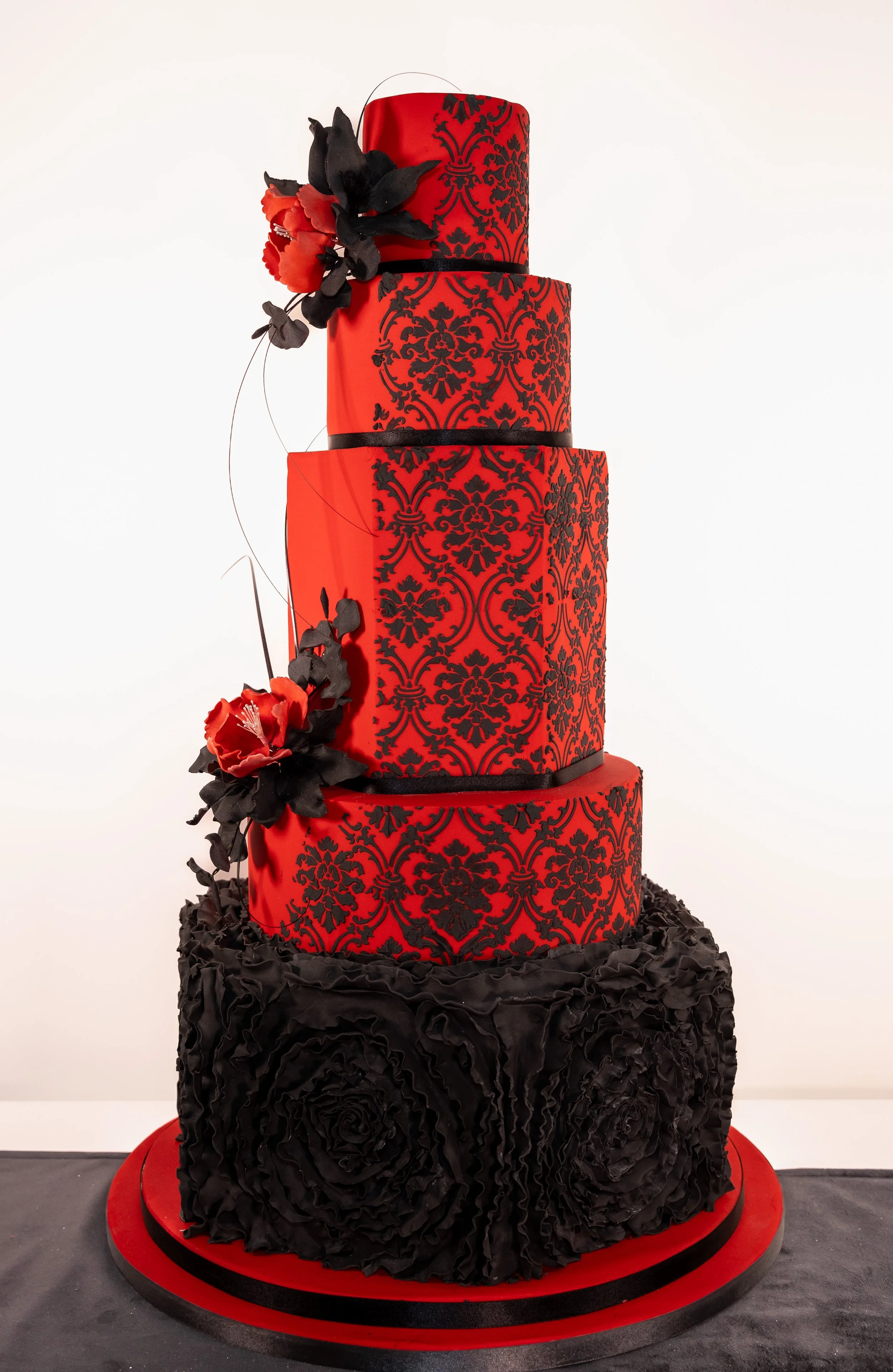 A four-tiered cake with black and red ornate designs, decorated with black and red flowers, sitting on a red and black round base.