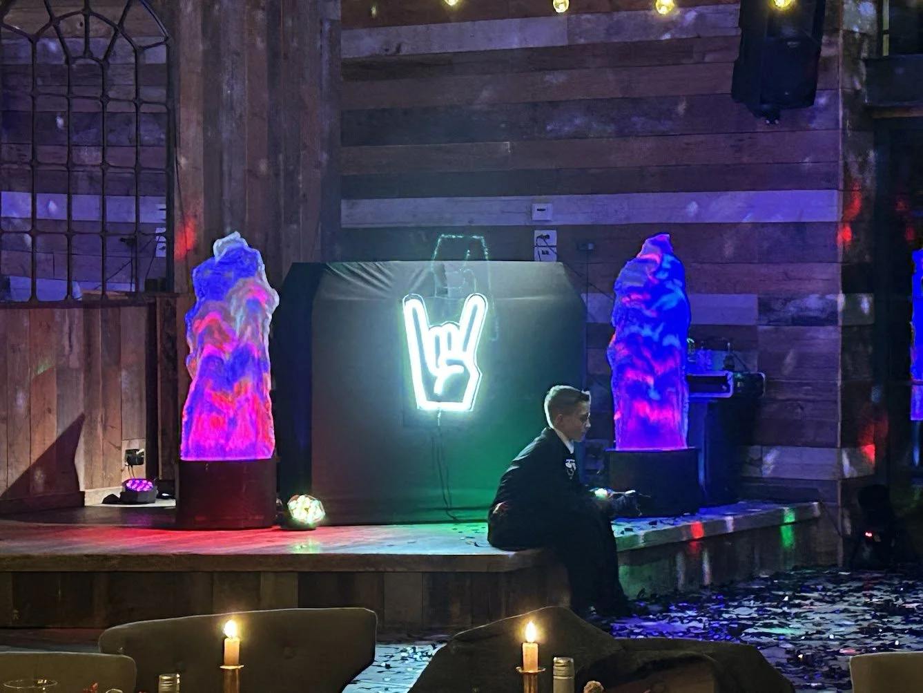 A young man sitting on a wooden stage in a dimly lit room decorated with colorful lights and neon signs, including a neon hand making the rock and roll sign, with two large abstract statues on either side of the stage and candles in the foreground.