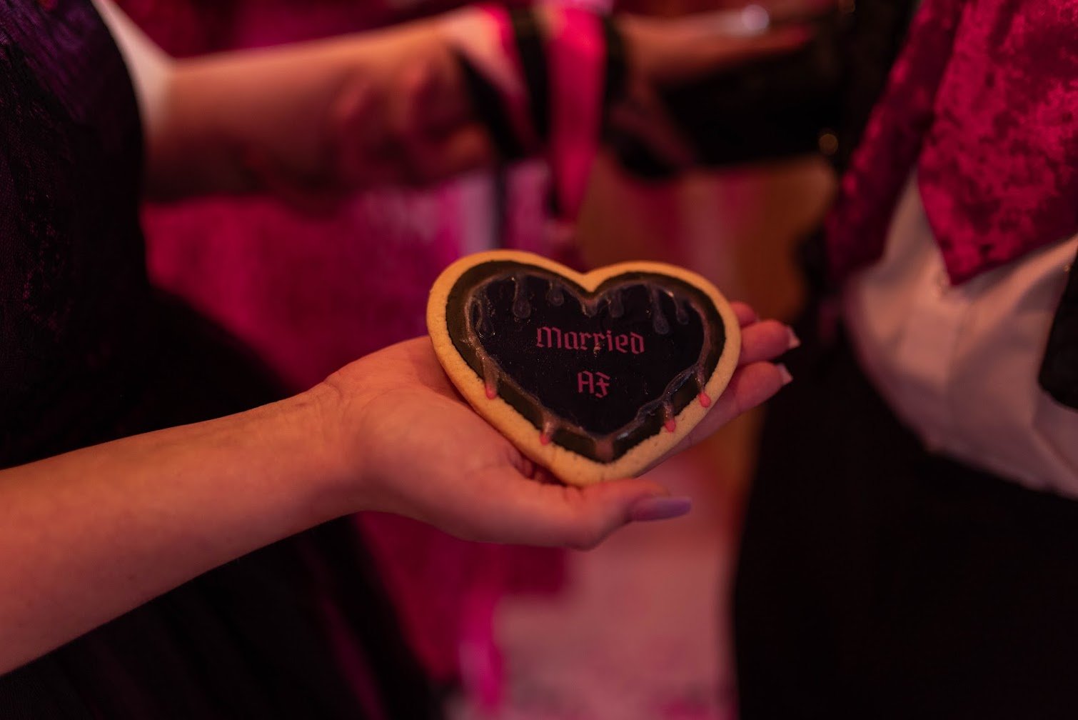 A person holding a heart-shaped cookie with black icing, on which 'Married' and initials 'AZ' are written in pink. The background shows other people at what appears to be a social or celebratory event.