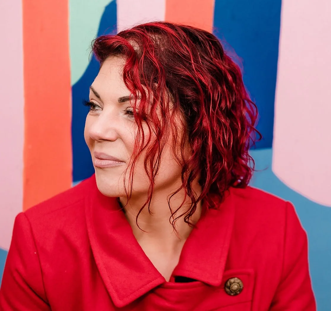 A woman with curly red hair wearing a red coat, looking to her right, standing in front of a colorful abstract background.