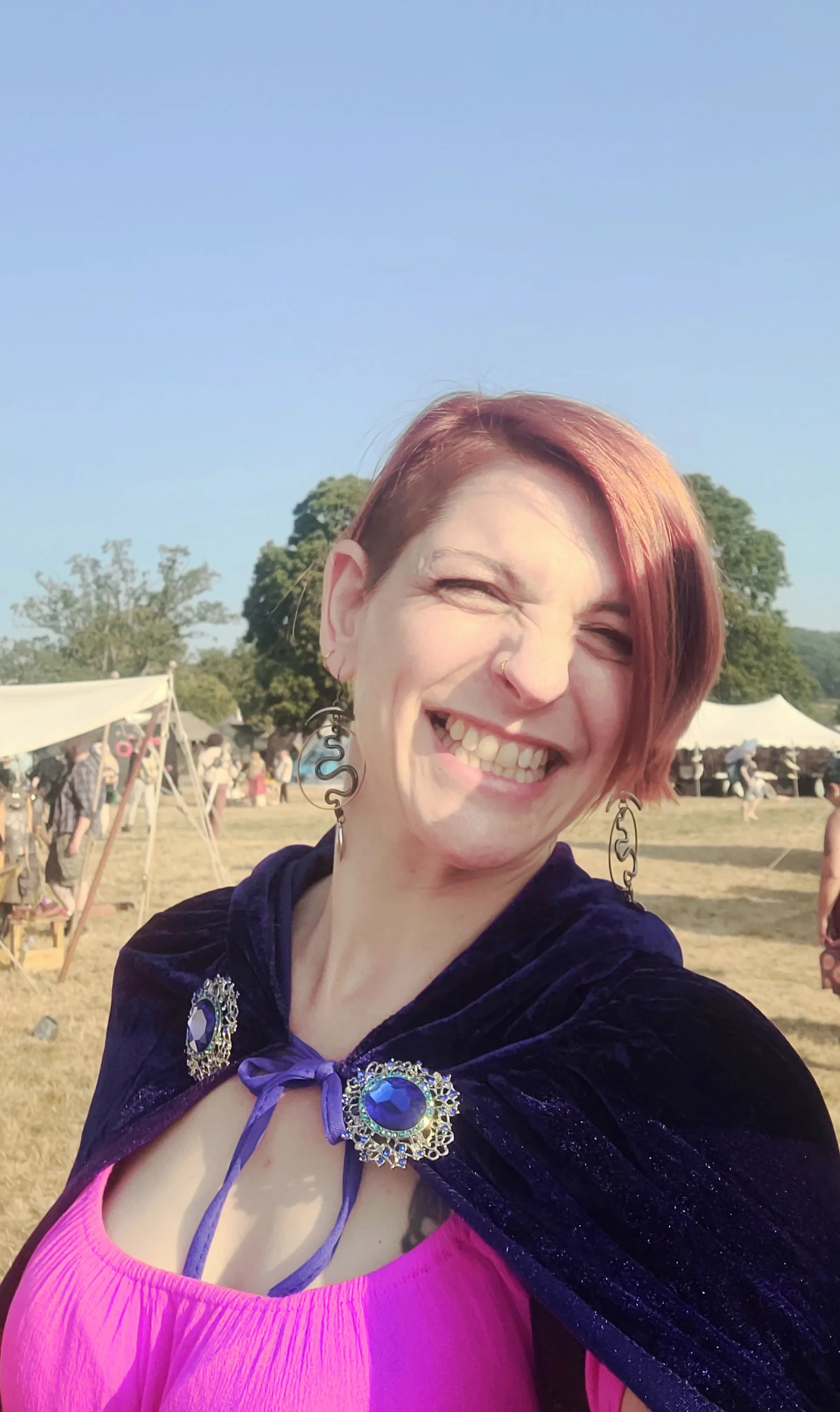 A woman smiling outdoors at a festival or fair, with tents and trees in the background. She has short red hair, is wearing a purple cape with ornate jewelry, and a bright pink top.
