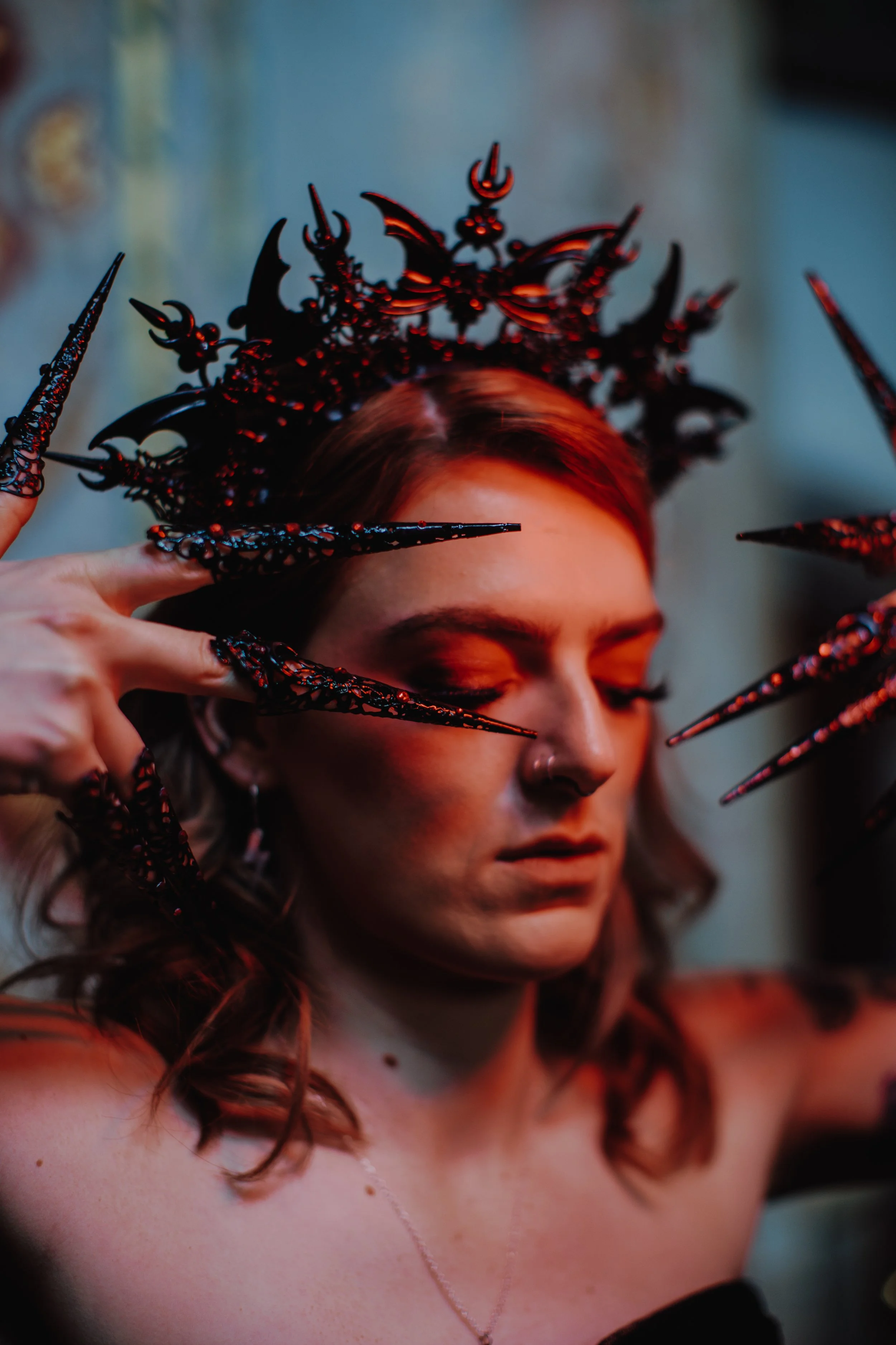 A woman with light skin and auburn hair wears elaborate black and red horns on her head and fiery red nails. She has her eyes closed and is posing with a serious expression, with her fingers near her face.