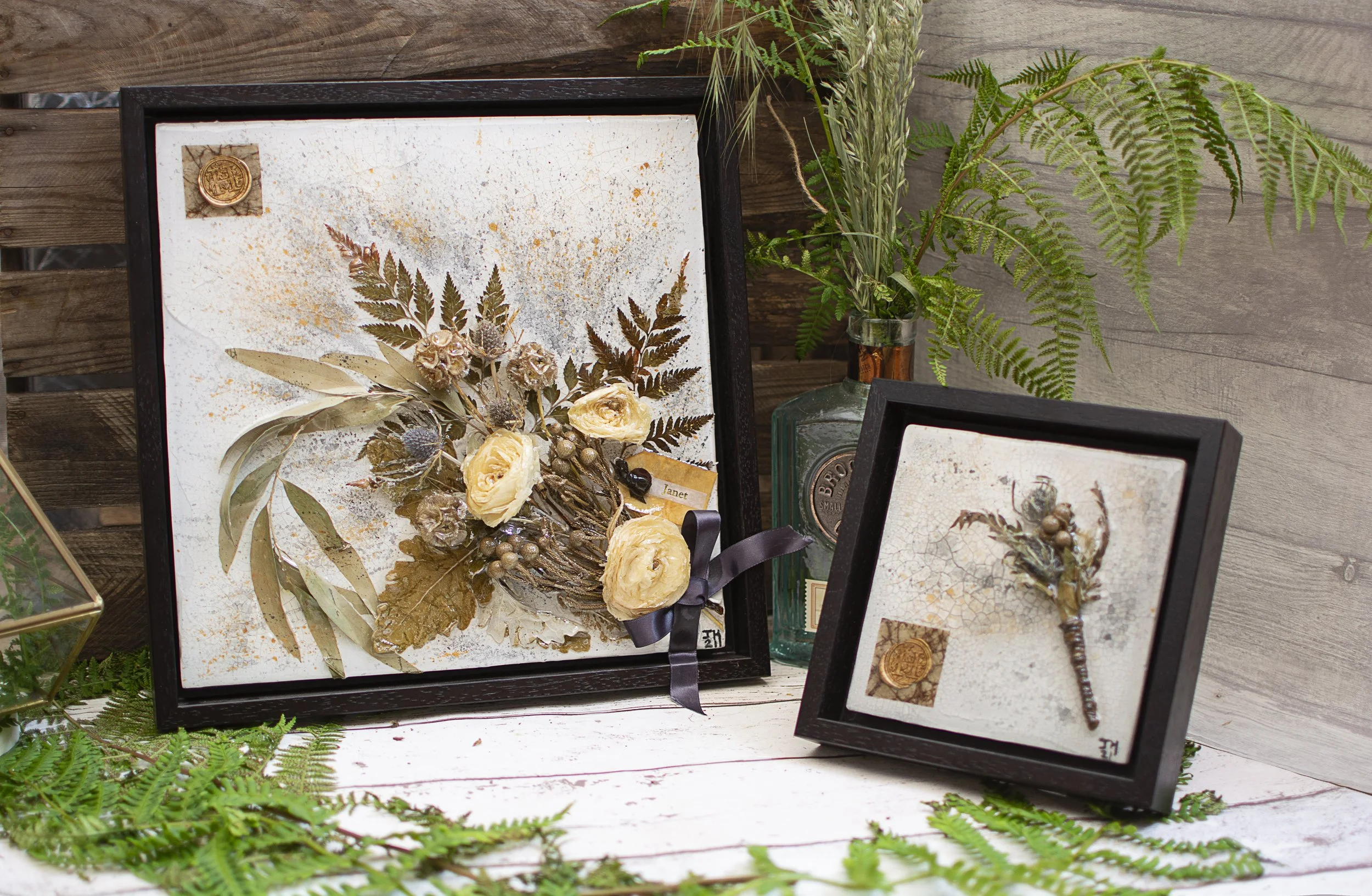Decorative framed artwork with dried flowers and foliage, a glass bottle with green leaves, and a wooden surface with greenery.