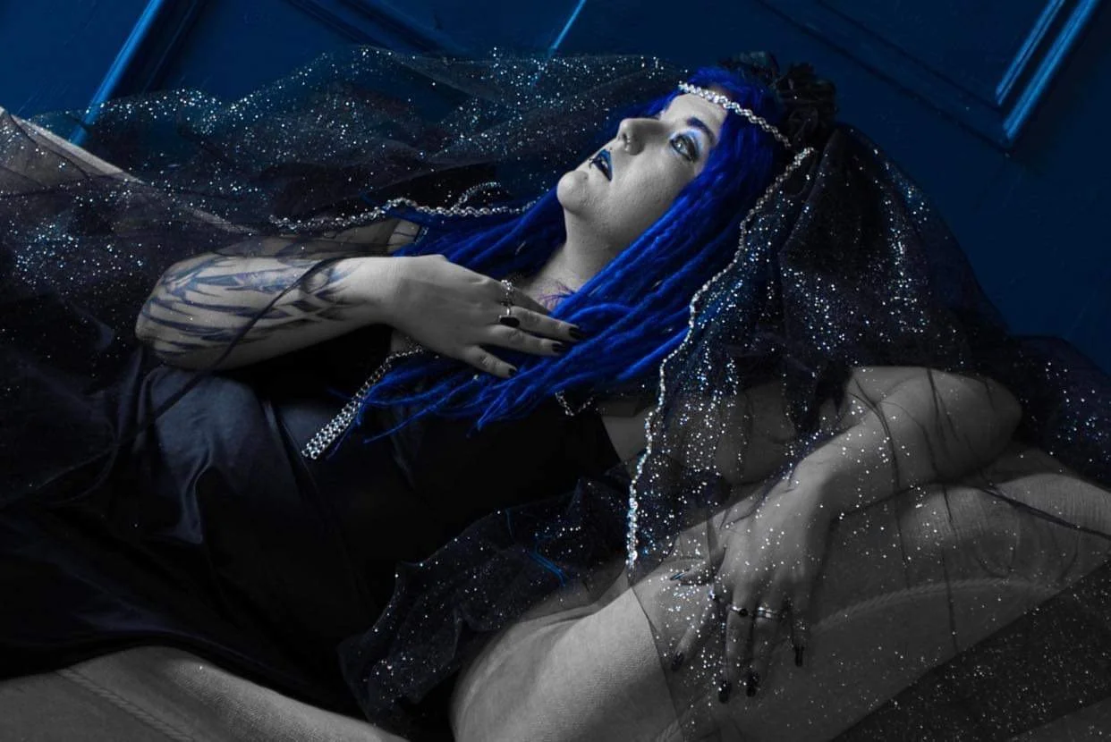 A woman with long blue dreadlocks lying on a sofa, wearing a black dress with metallic accents and a sheer, glittery, starry cape, in a dark, moody setting.