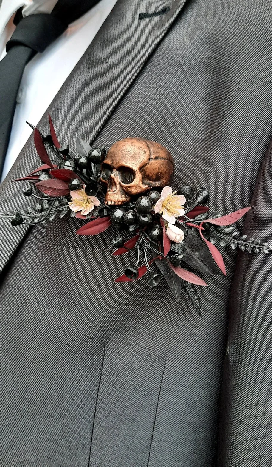 A dark-colored suit jacket with a skull and floral pin on the lapel, featuring a small skull, black berries, pink flowers, and dark red leaves.