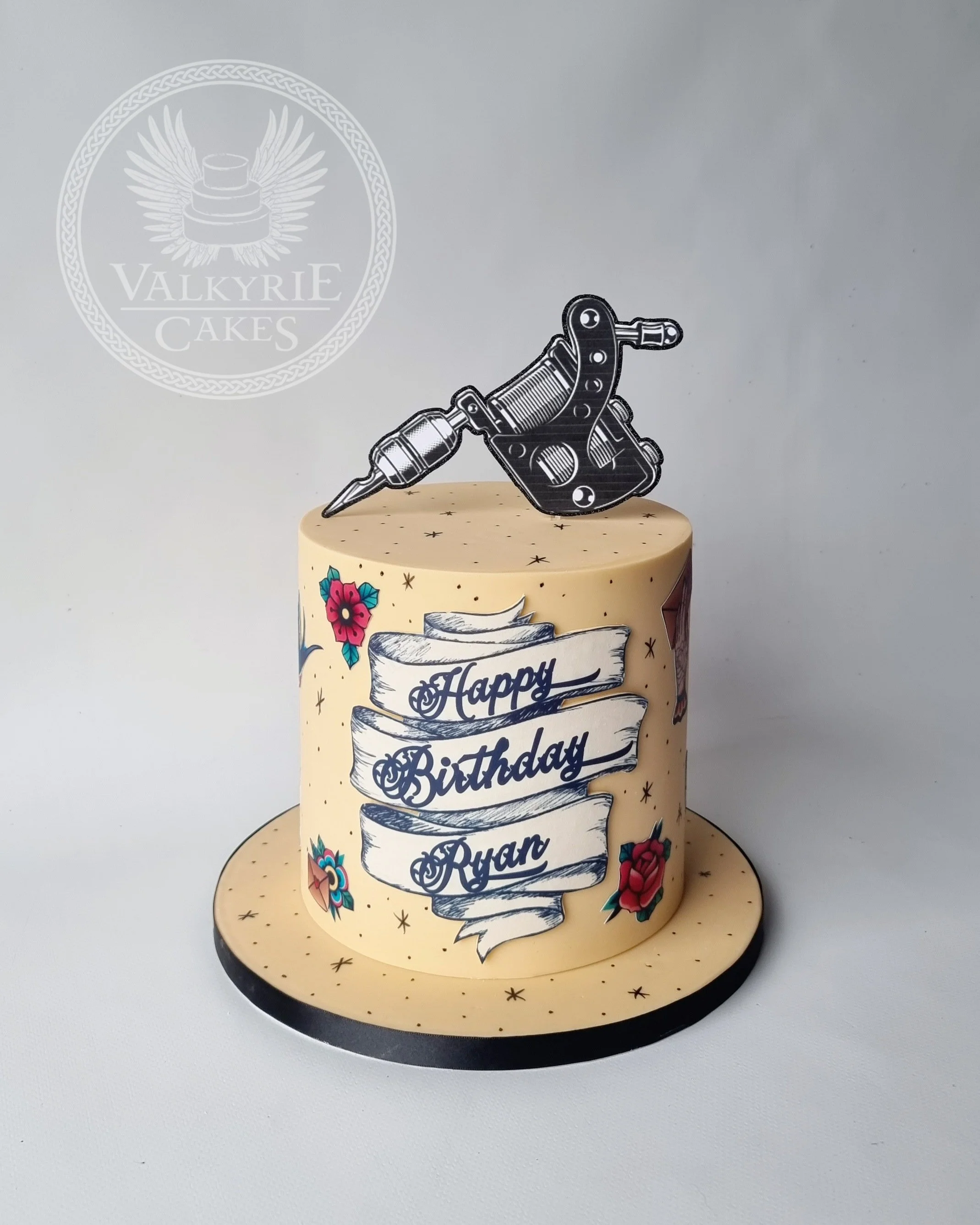 A birthday cake with yellow icing and tattoo-style flower decorations, topped with a tattoo machine and syringe cake topper, and a 'Happy Birthday Ryan' message written on a banner.