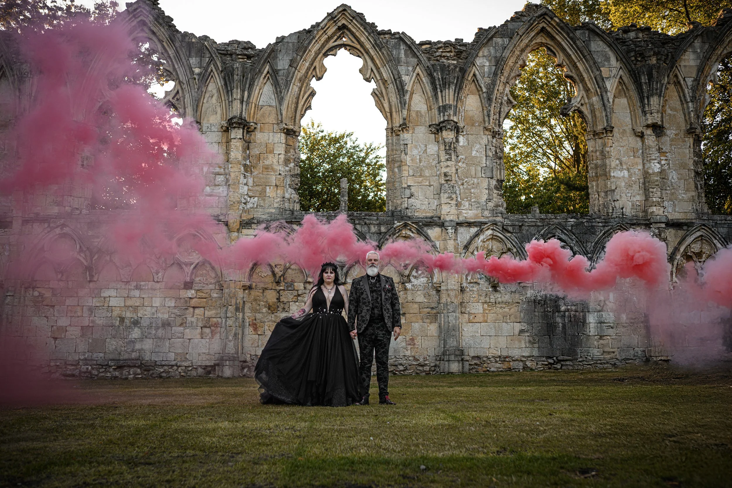 A couple dressed in gothic attire standing in front of ancient stone ruins, holding hands, with pink smoke billowing across the scene.
