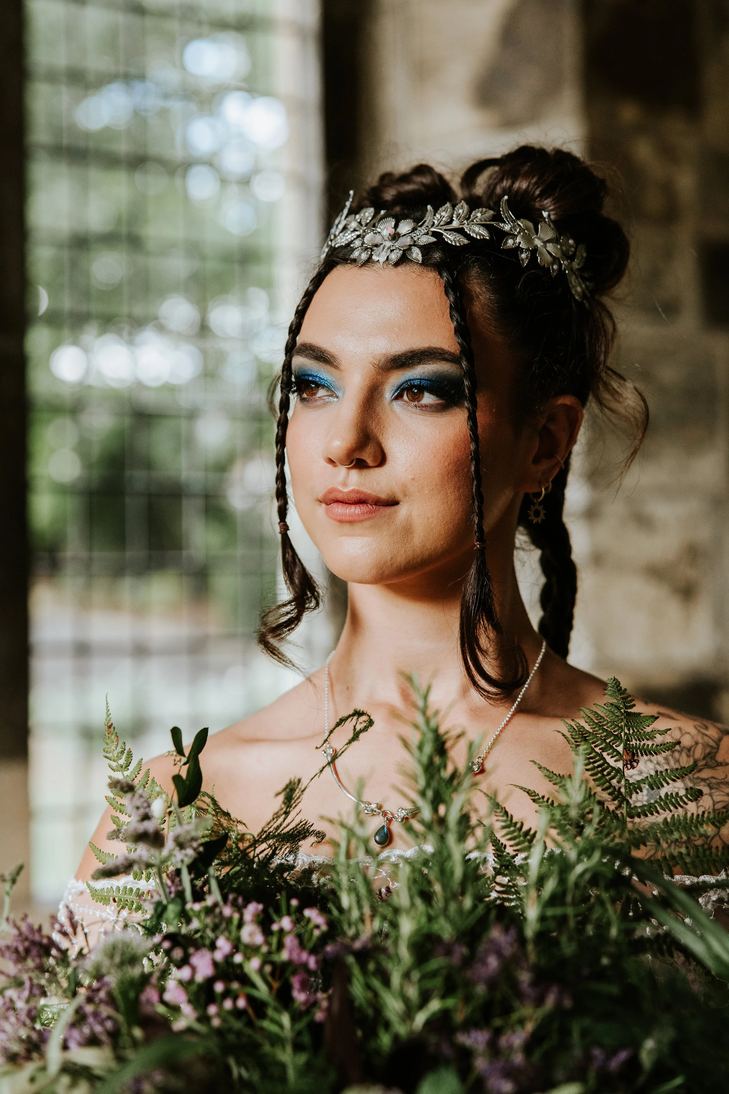 A woman with dark hair styled in updo with braids, wearing a silver leaf crown, blue eye makeup, and earrings, holding a bouquet of greenery and flowers, in a rustic setting.