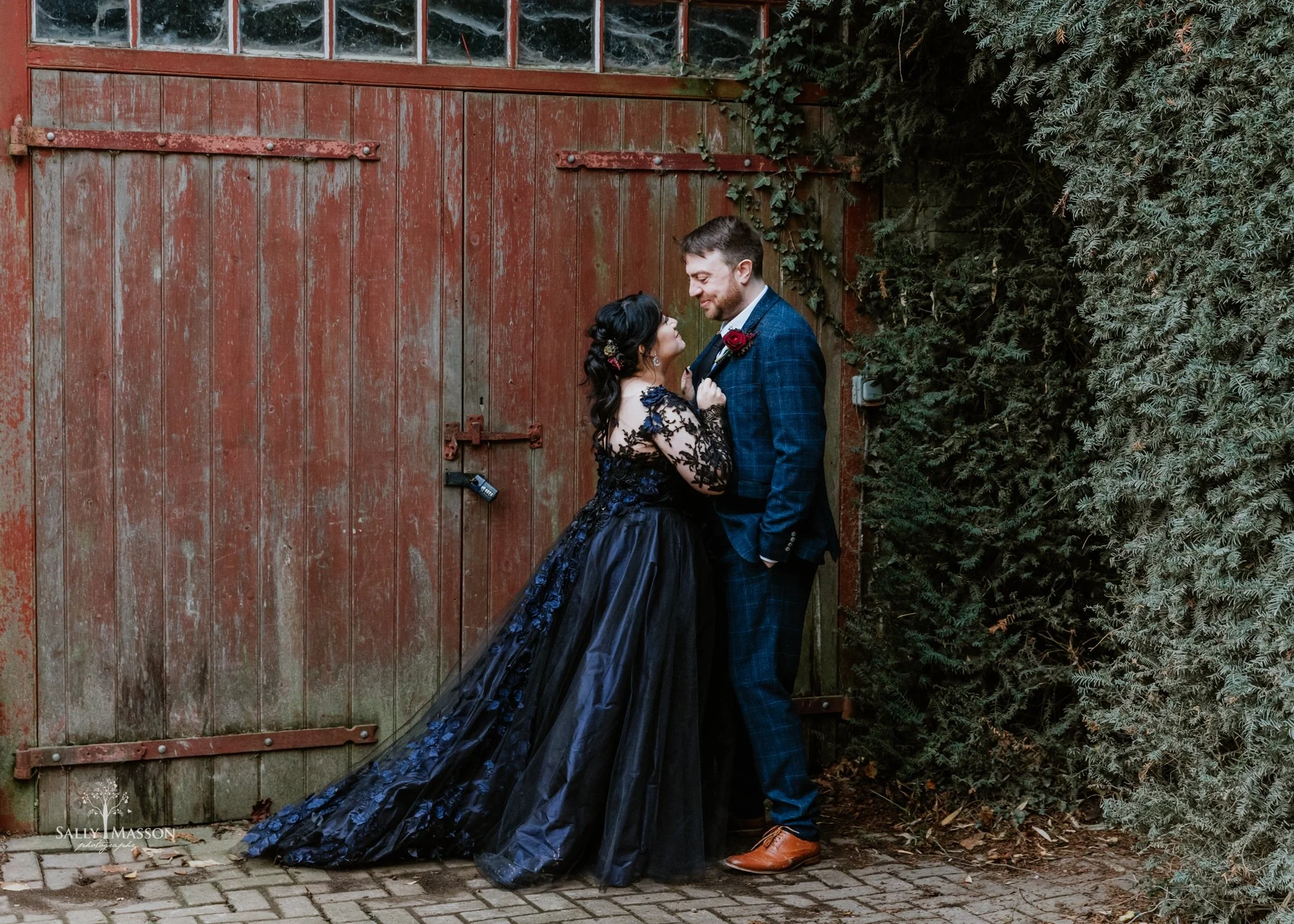 A couple dressed in wedding attire standing in front of a rustic red wooden gate and green foliage. The woman wears a black, lace, long-sleeved gown with a train, and the man wears a dark blue plaid suit with a red flower boutonniere. They are looking at each other affectionately.