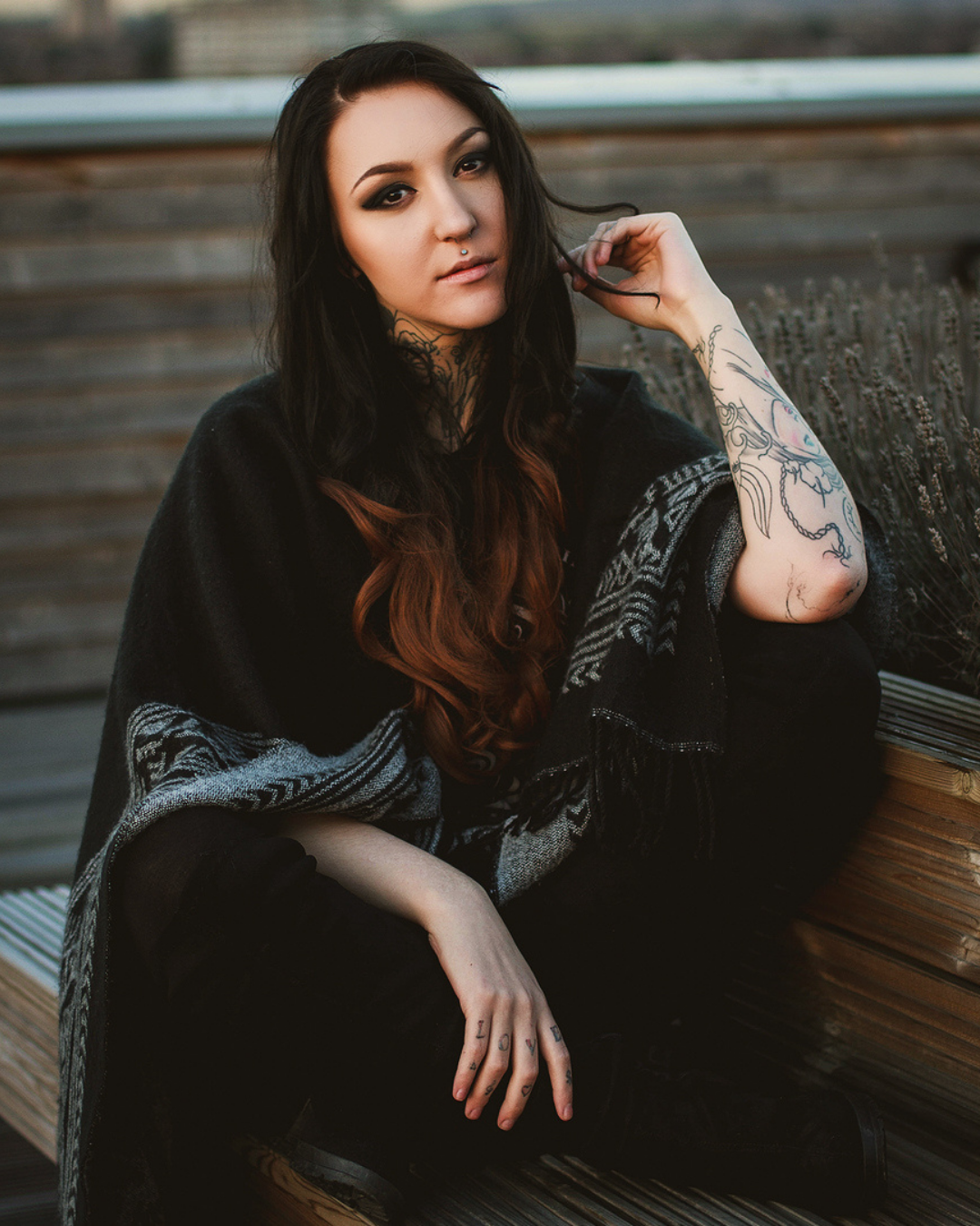 A young woman with long dark hair, tattoos, and piercings sitting on a wooden bench outdoors.