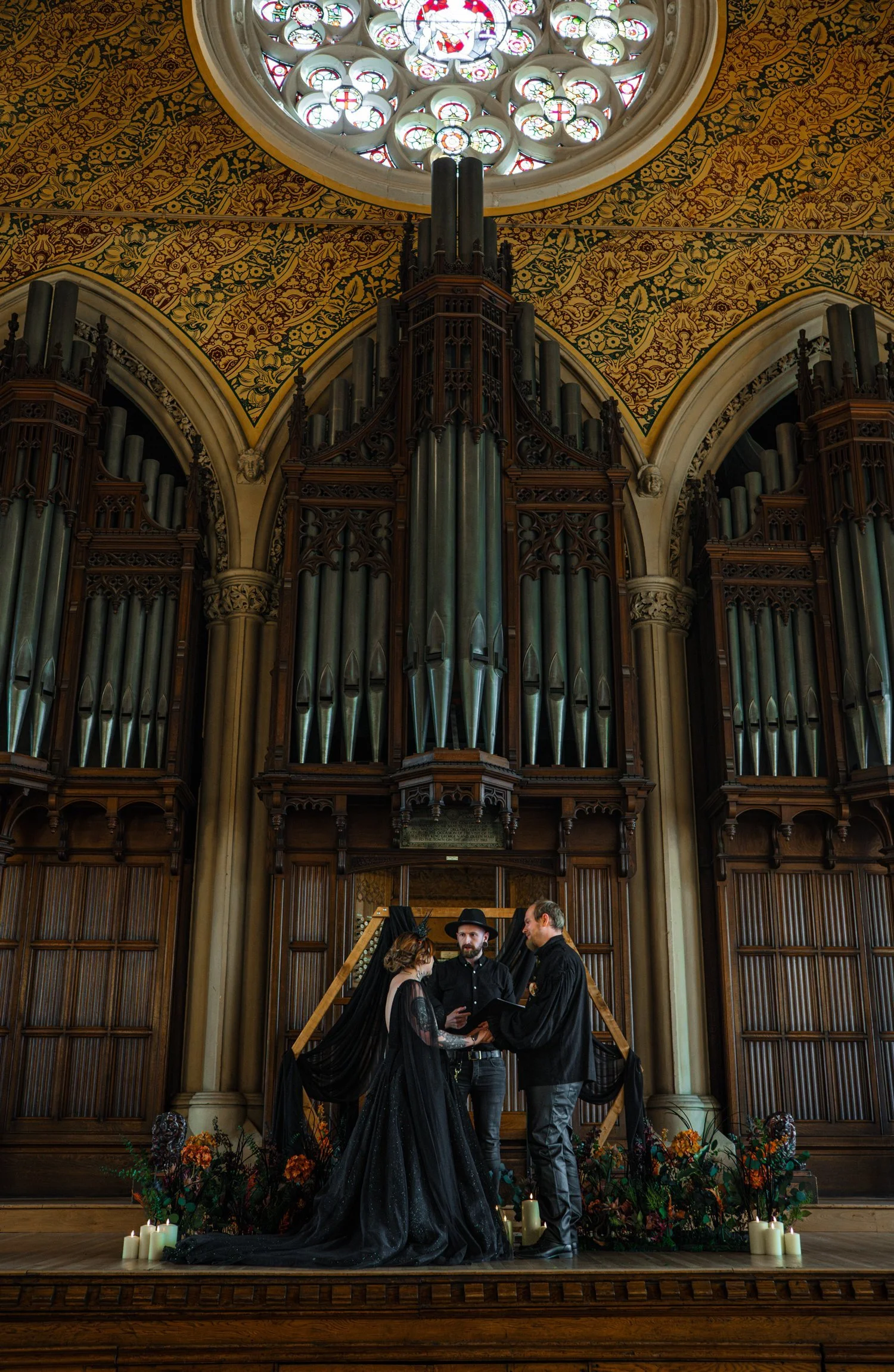 A couple dressed in gothic style wedding attire standing before a person officiating their wedding in front of a large pipe organ in a church with stained glass ceiling and ornate interior.