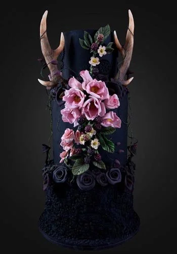 A decorative headpiece with antlers, pink roses, purple roses, white daisies, green leaves, and black lace on a dark background.