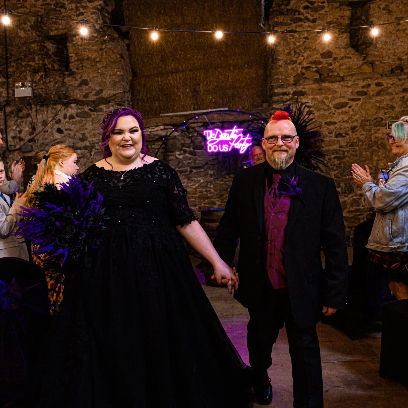 A woman with purple hair in a black dress holding a purple bouquet, and a man with a mohawk hairstyle with red hair, mustache, and beard wearing a black suit with a purple shirt, are holding hands and smiling at an event with a rustic stone wall and purple neon sign in the background.